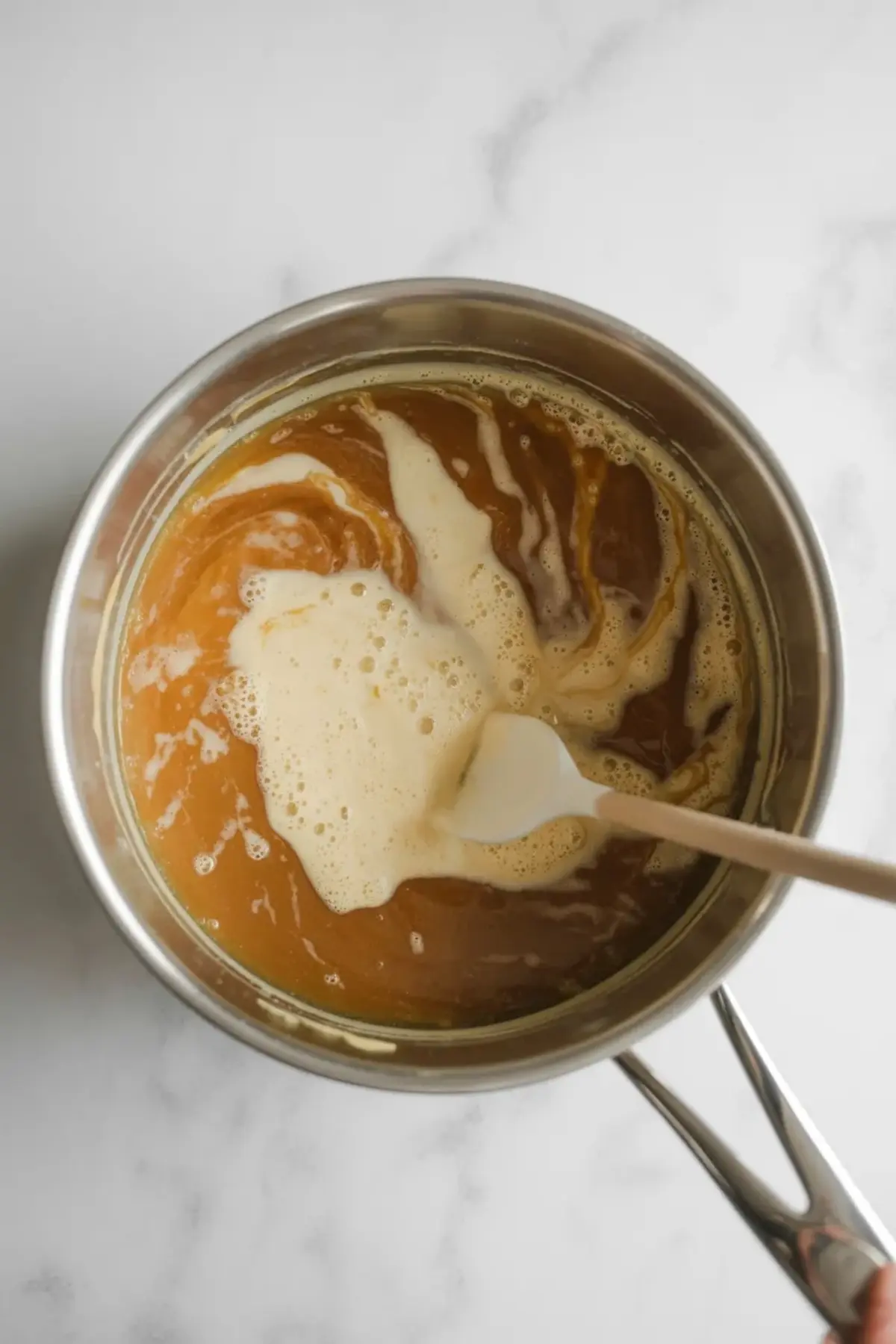 A saucepan with rich caramel sauce in the process of blending, with a white spatula stirring in cream, creating a marbled texture on the surface.
