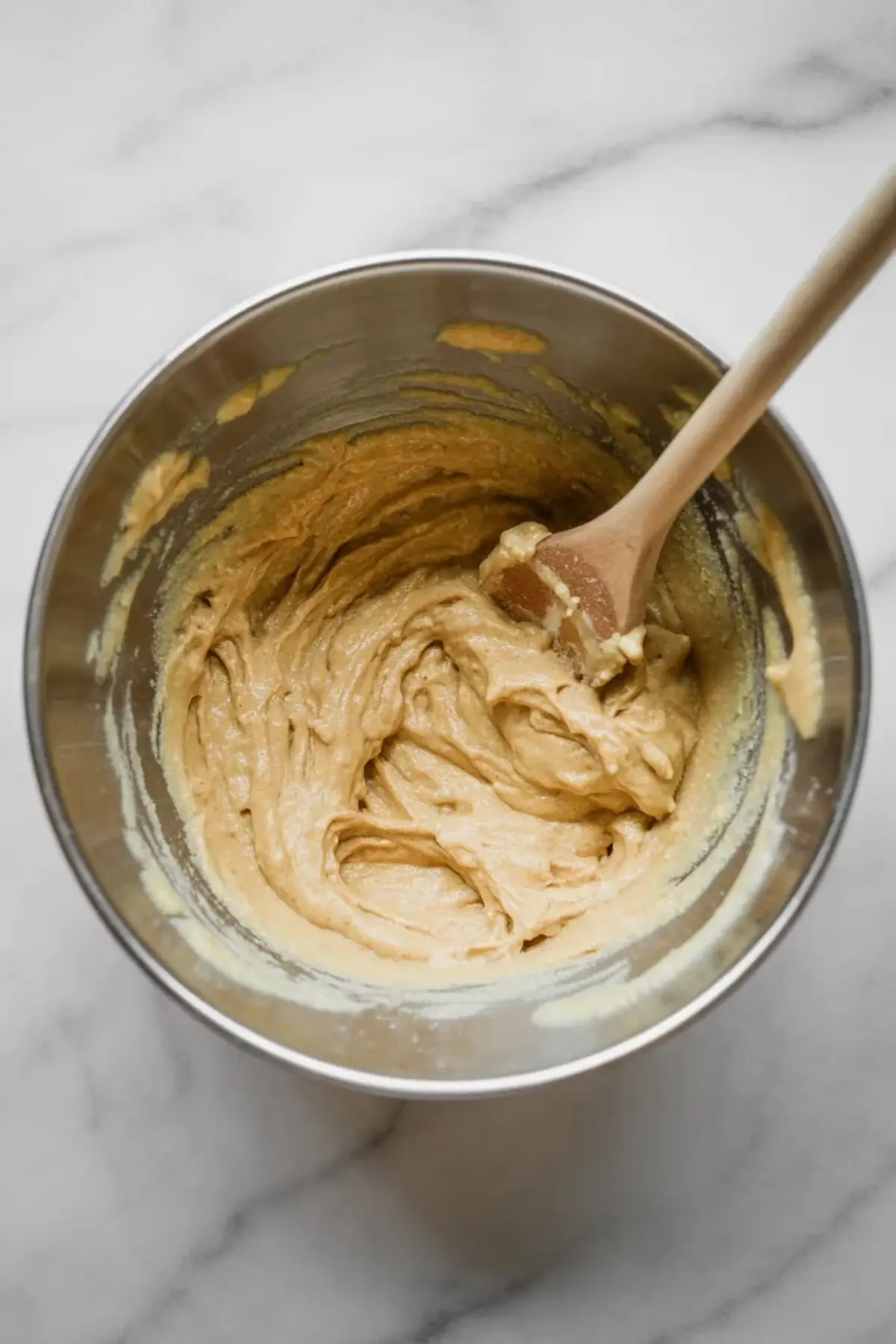 Creamy muffin batter mixed until smooth in a stainless steel bowl with a wooden spoon resting inside, placed on a light marble countertop.