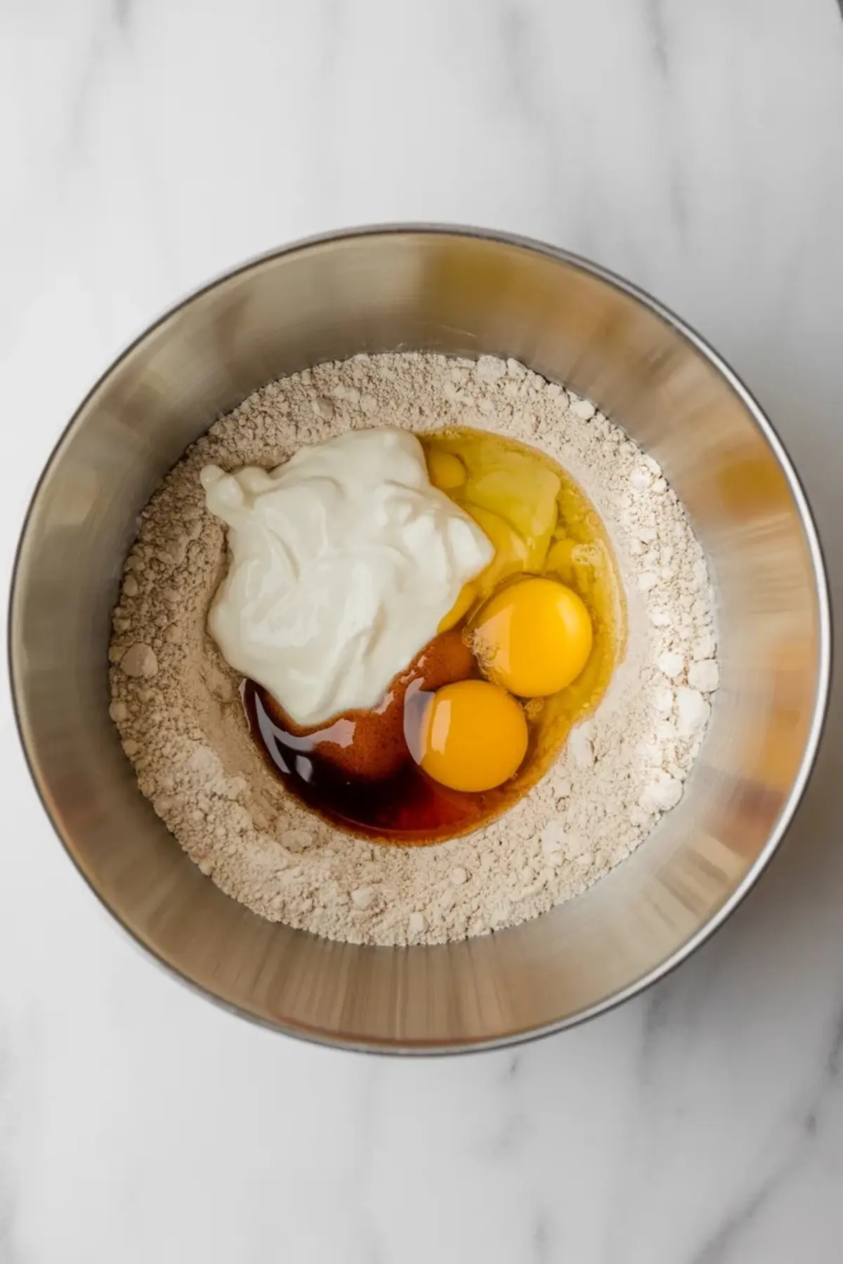 Baking ingredients in a stainless steel bowl including eggs, sour cream, vanilla extract, and flour, shown before mixing on a white marble background.
