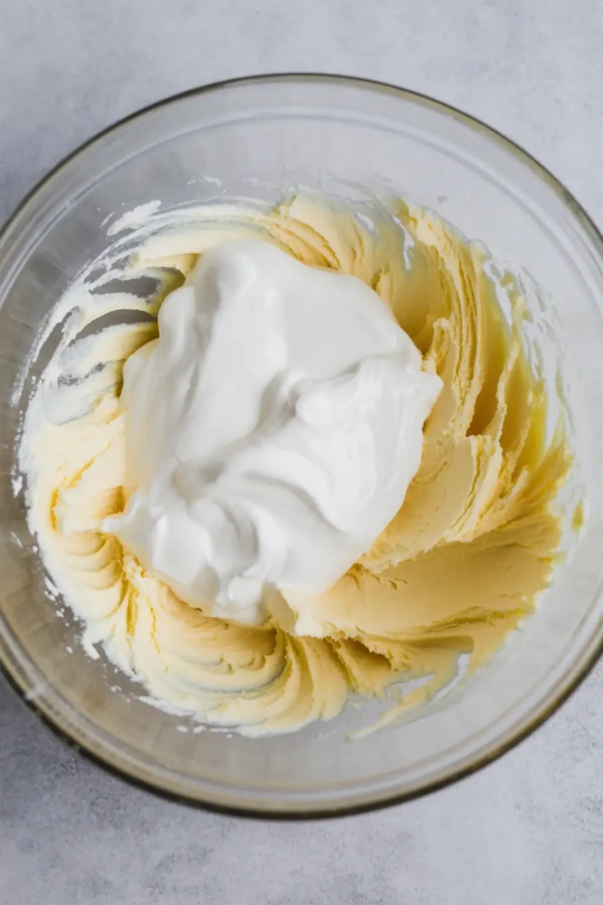 Partially mixed sour cream frosting in a glass bowl, combining whipped cream with creamed butter and sugar for a thick and airy consistency.
