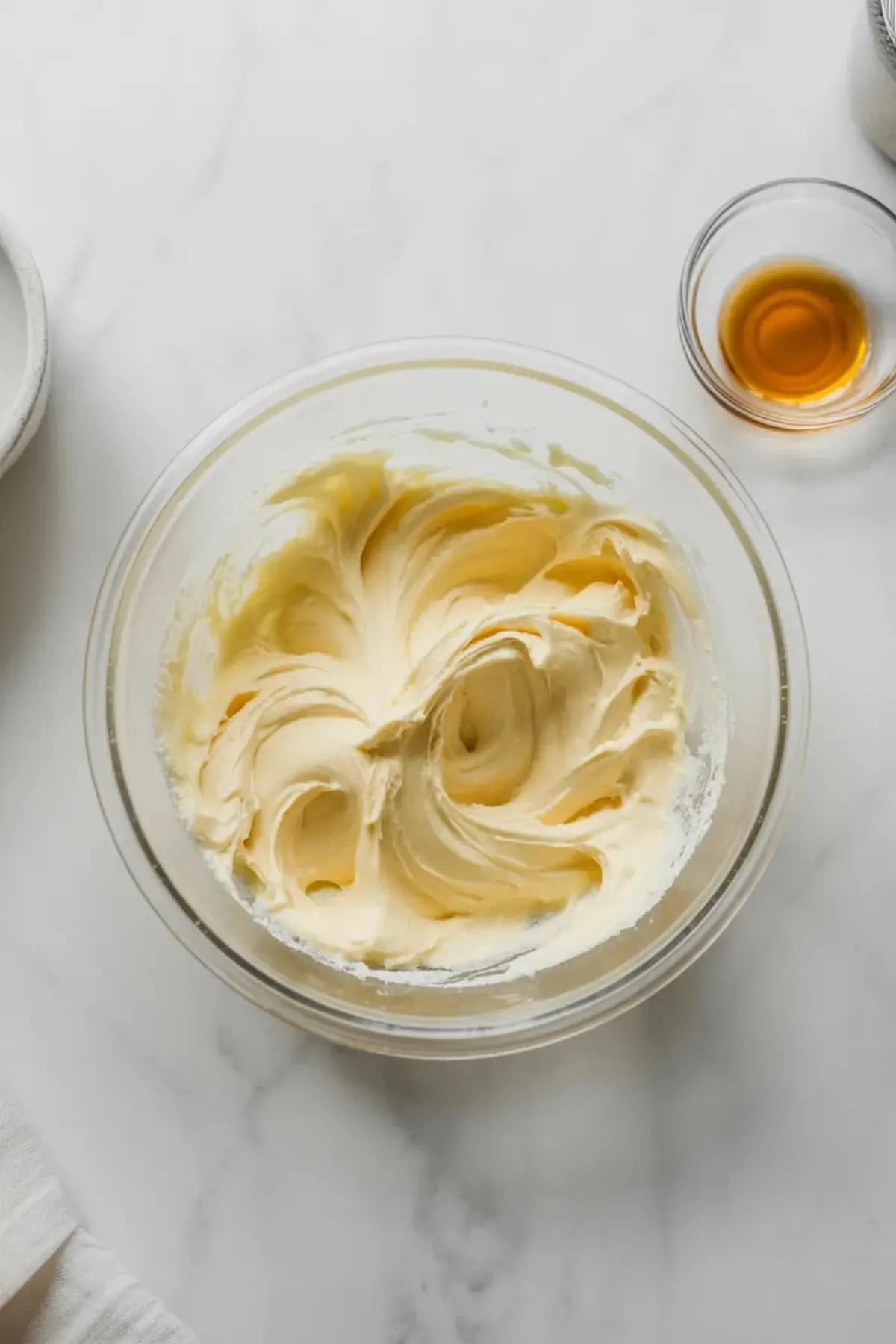 Whipped sour cream frosting in a glass bowl with soft peaks and smooth texture, placed beside a small bowl of vanilla extract on a white countertop.
