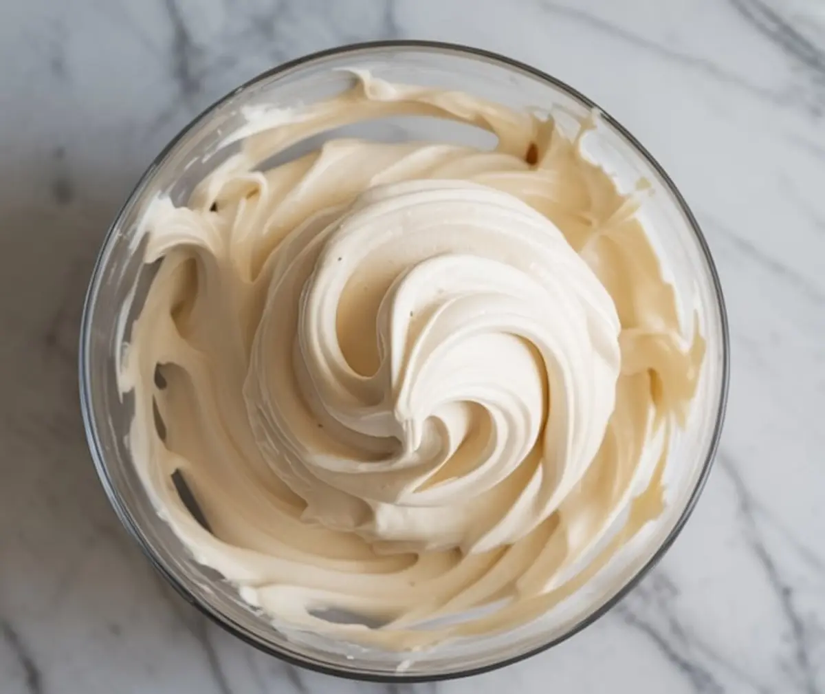 Lightly golden Swiss meringue buttercream piped in a tall swirl inside a clear glass bowl, placed on a marble surface with soft shadows and creamy texture.