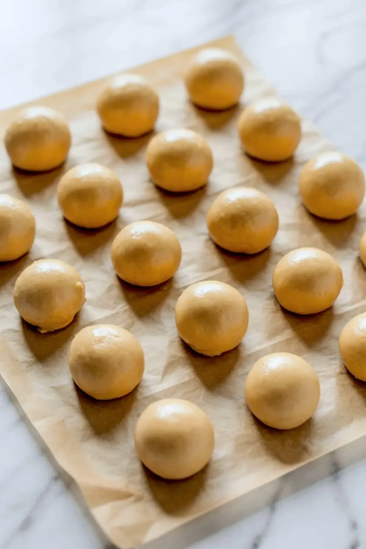 Round balls of cookie dough are arranged in neat rows on a parchment-lined baking sheet, ready to be baked for Twix thumbprint cookies.