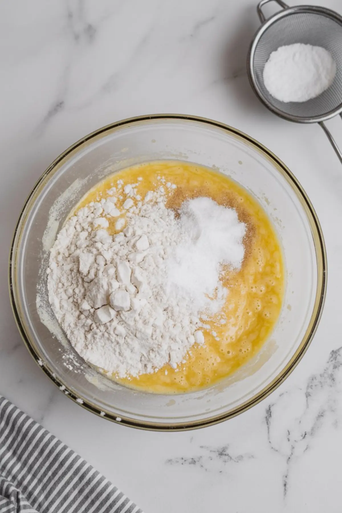 Glass mixing bowl filled with whisked wet ingredients, white flour, baking soda, and salt, ready for mixing into cookie dough, staged on a marble surface with a mesh strainer nearby.
