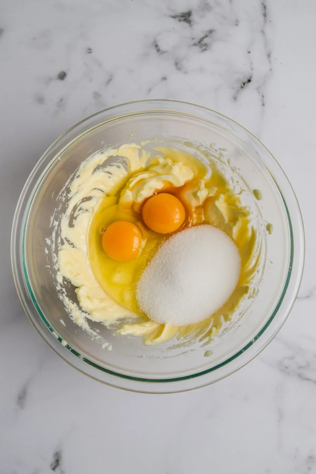 Glass mixing bowl with creamed butter, two egg yolks, vanilla extract, and granulated sugar on a marble surface, showing wet ingredients for making a cookie skillet dough.
