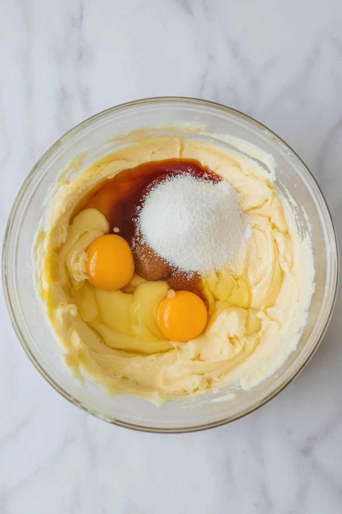 Creamed butter and sugar mixture in a glass bowl with cracked eggs, vanilla extract, and white granulated sugar added on top.
