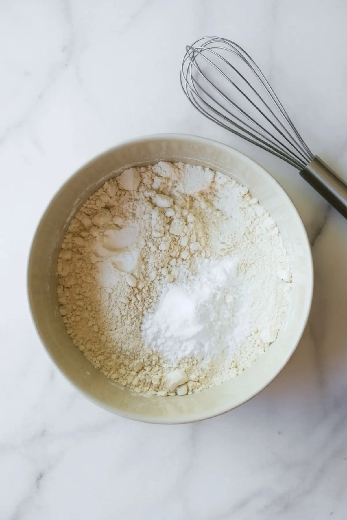Mixing bowl filled with flour, baking powder, and baking soda blend, with a metal whisk beside it on a white marble surface.
