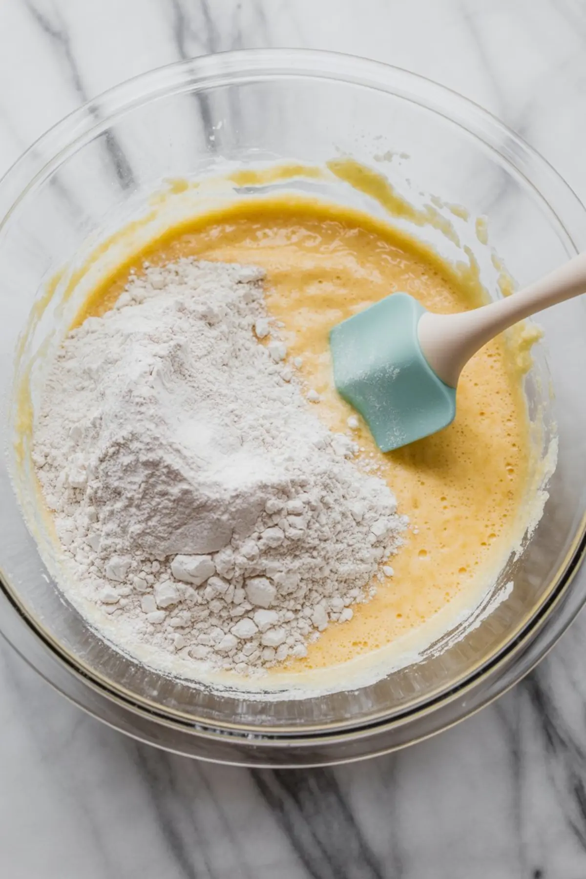 Glass bowl shows flour folded into yellow cake batter with a silicone spatula, capturing the mixing step for homemade coconut cake batter.