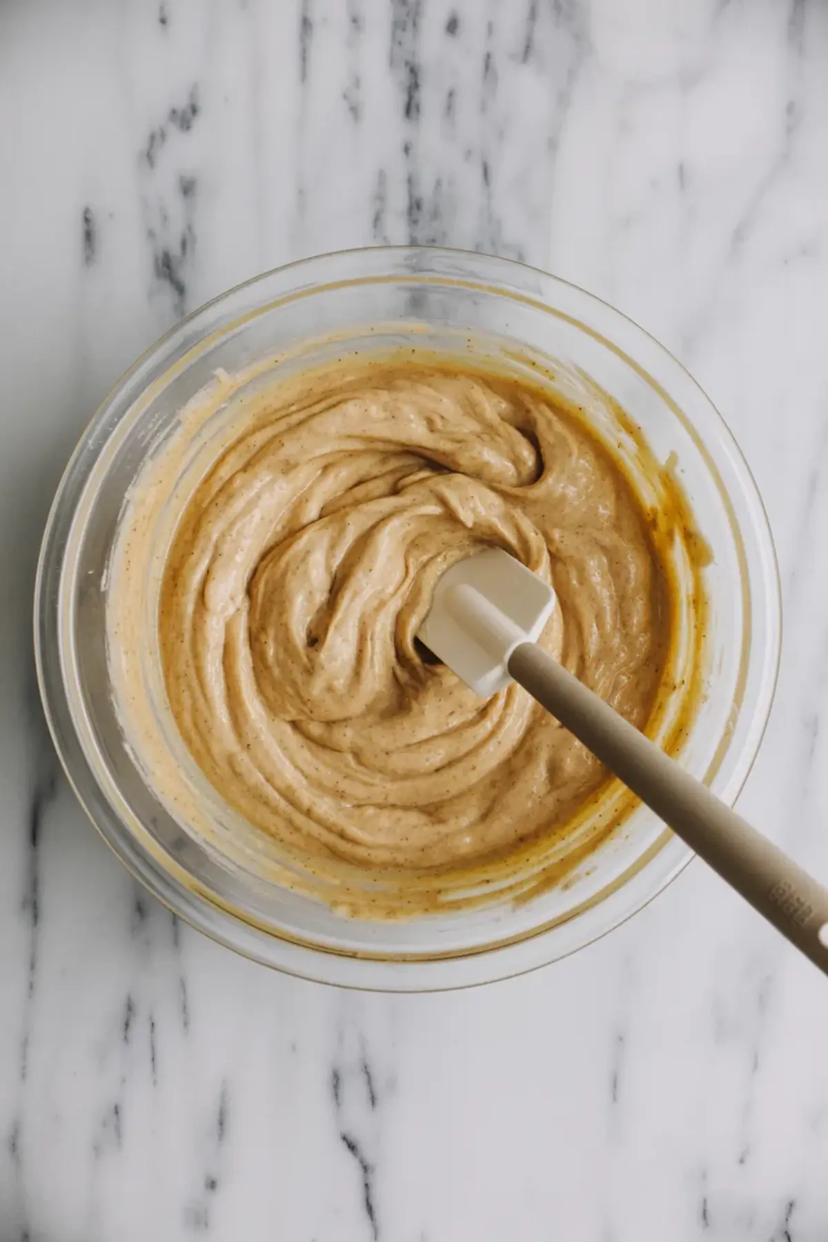 Creamy almond flour banana bread batter swirled in a glass mixing bowl with a spatula on a marble surface, showcasing a smooth blend of mashed banana and almond flour.
