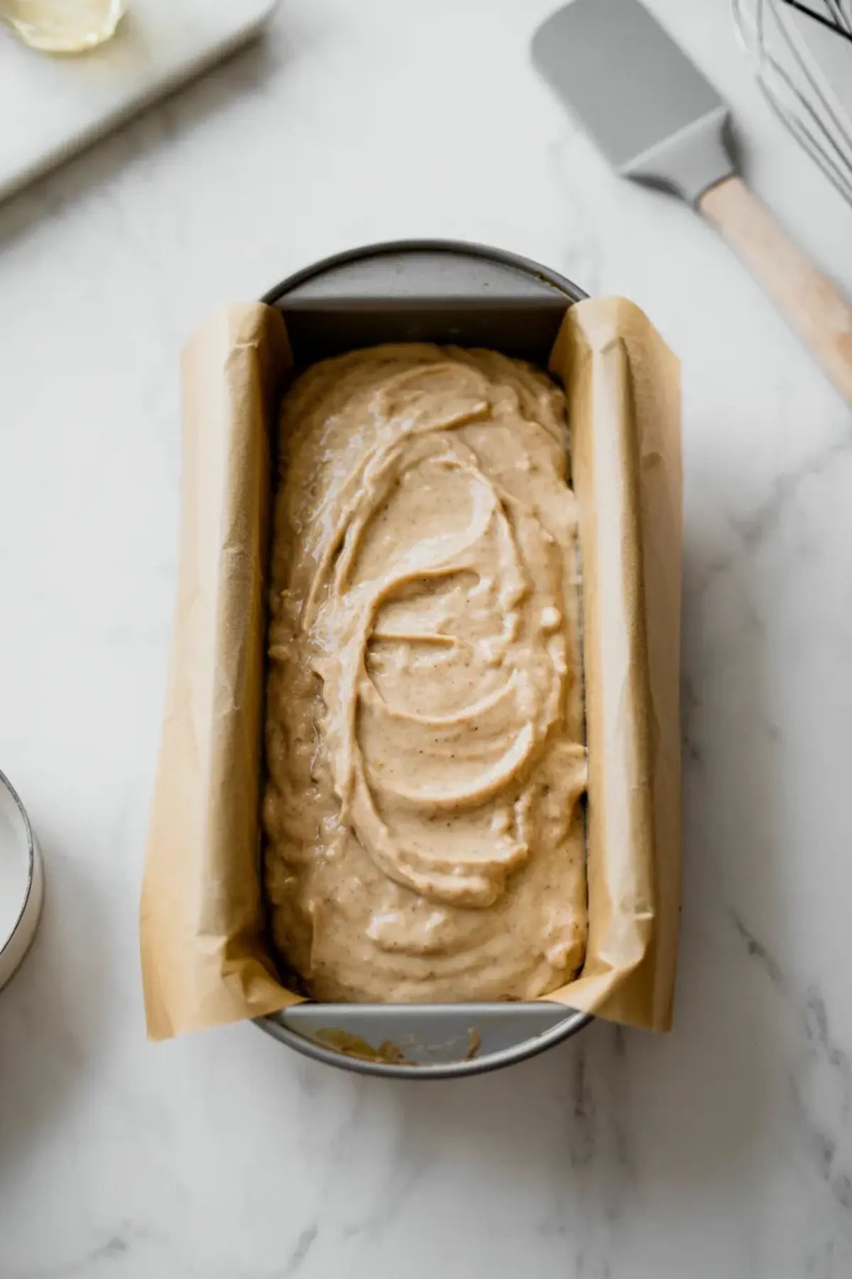 Almond flour banana bread batter evenly spread in a parchment-lined loaf pan, ready for baking on a white marble countertop with baking tools nearby.
