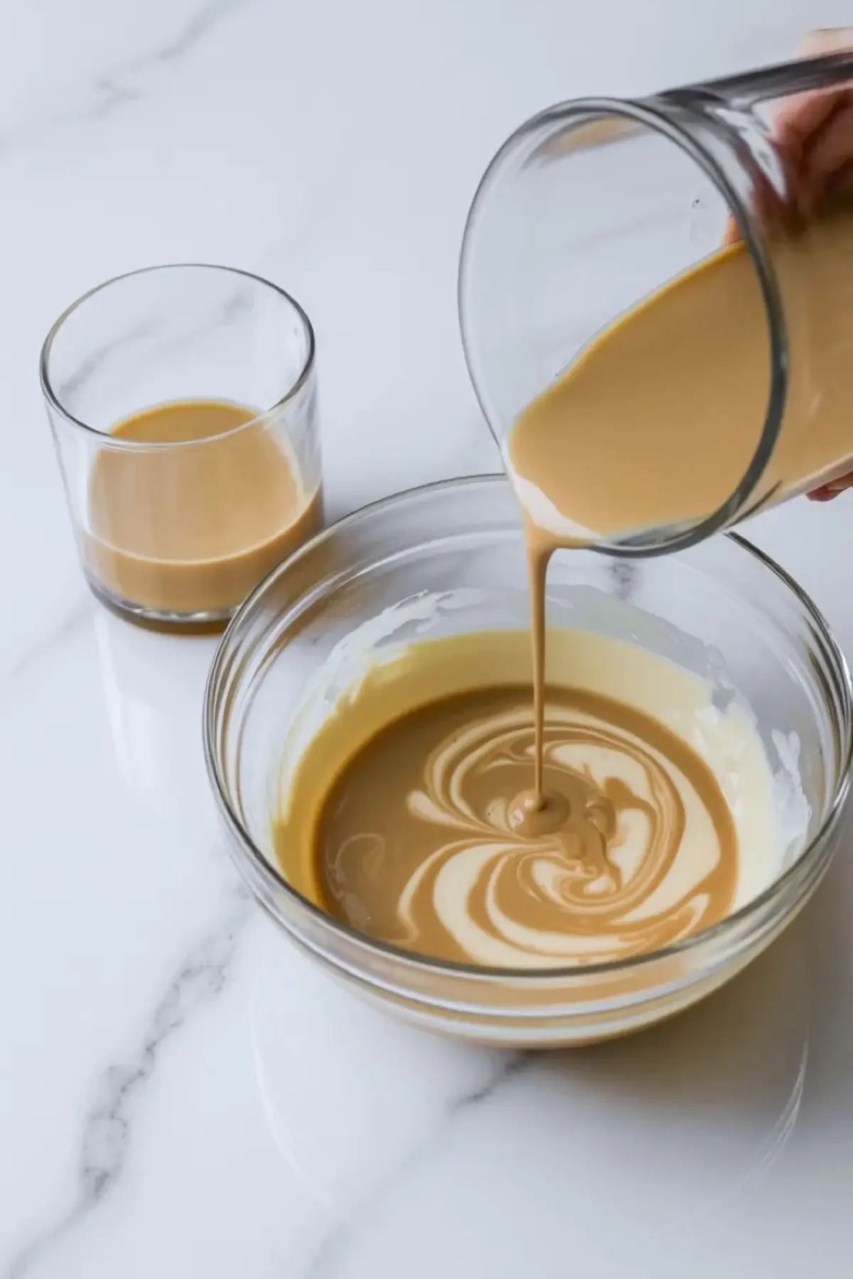 Baileys Irish Cream being poured into a bowl of cream mixture, creating a smooth swirl with a glass of liqueur in the background on a white surface.
