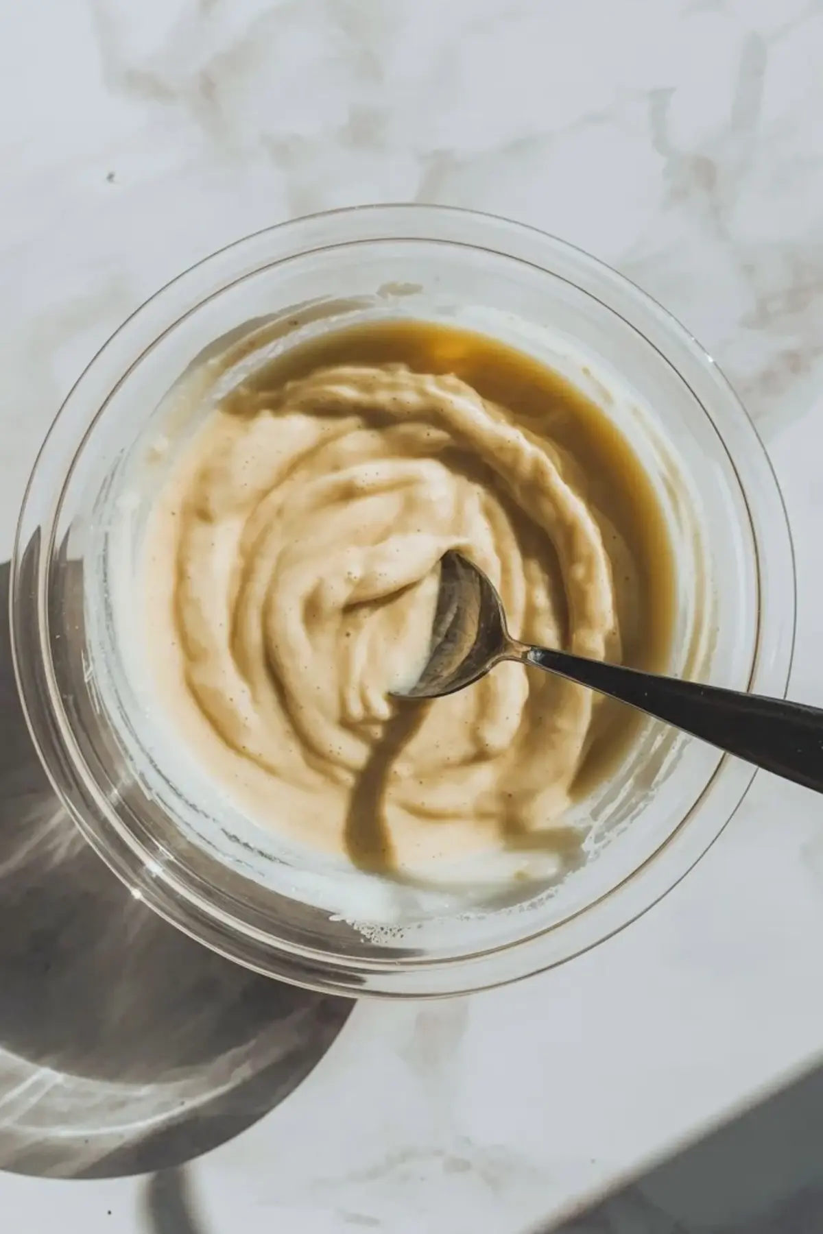 Top view of a glass bowl filled with smooth mashed banana mixture, a spoon rests inside the creamy banana puree on a light marble countertop for a healthy pudding base.