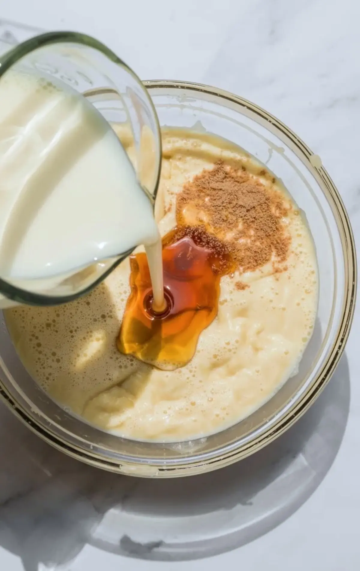 Milk pours from a glass into mashed banana mixture in a bowl, honey and ground cinnamon rest on top, showing simple ingredients for homemade banana chia pudding.