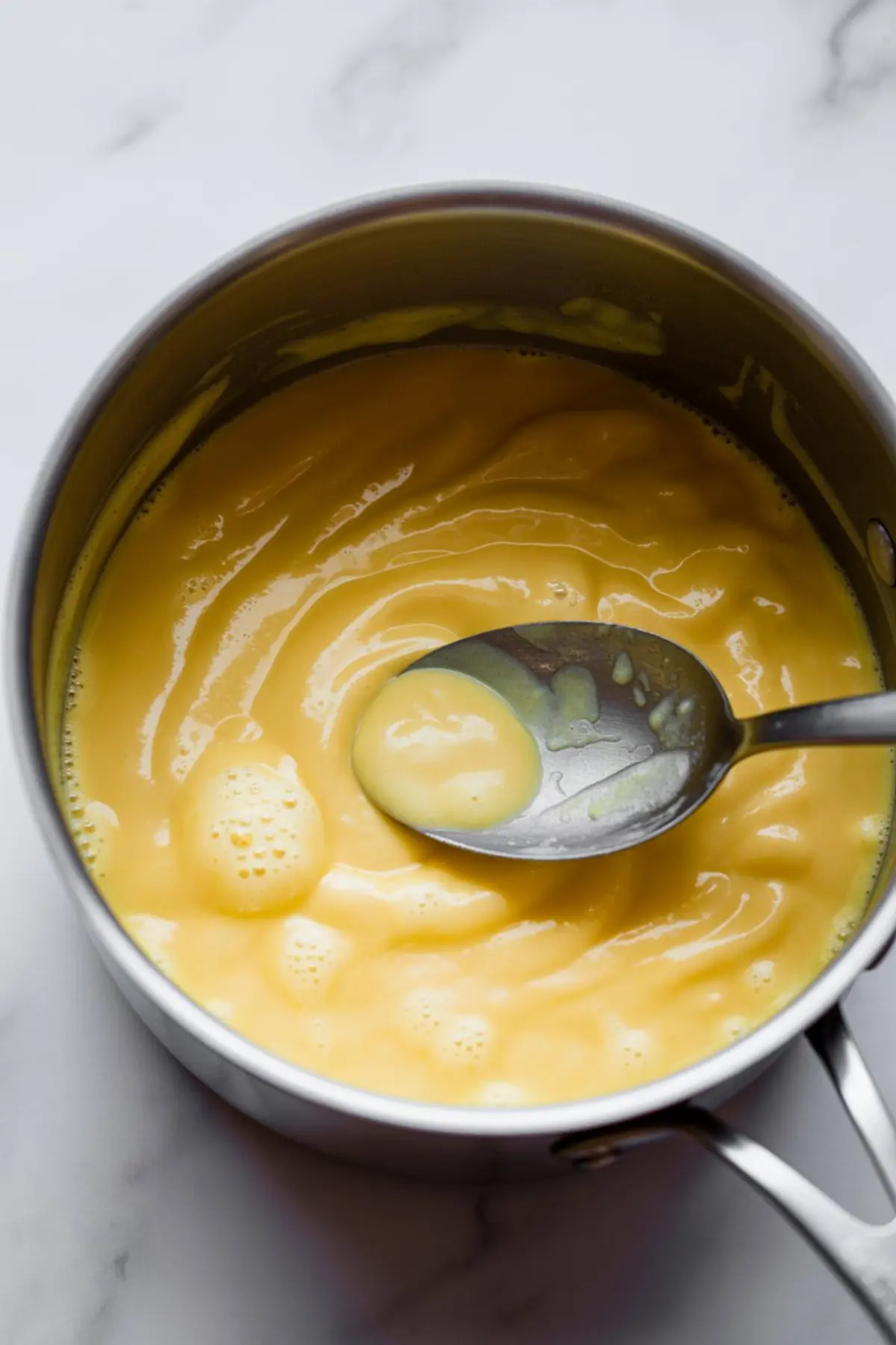 Saucepan filled with thick yellow custard being stirred with a spoon over a marble counter.
