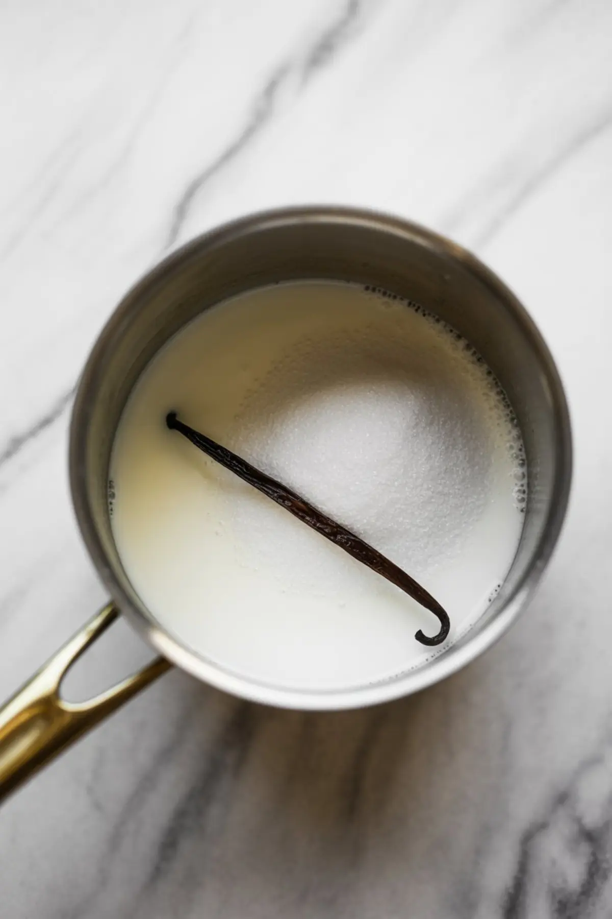 Saucepan containing milk, granulated sugar, and a vanilla bean pod resting on the surface.
