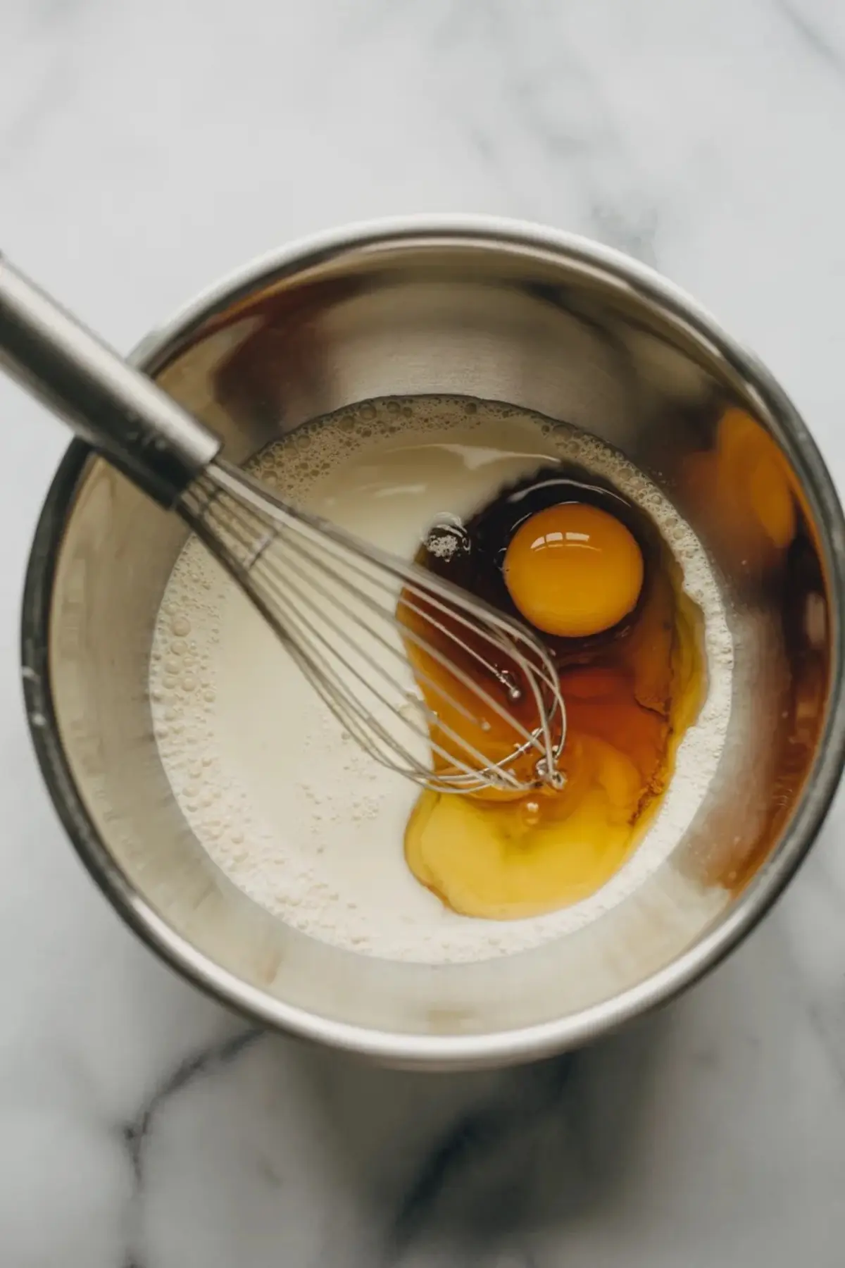 Egg, milk, maple syrup, and vanilla extract sit in a metal mixing bowl with a whisk. Wet ingredients combine to form batter for blueberry baked oatmeal breakfast casserole.
