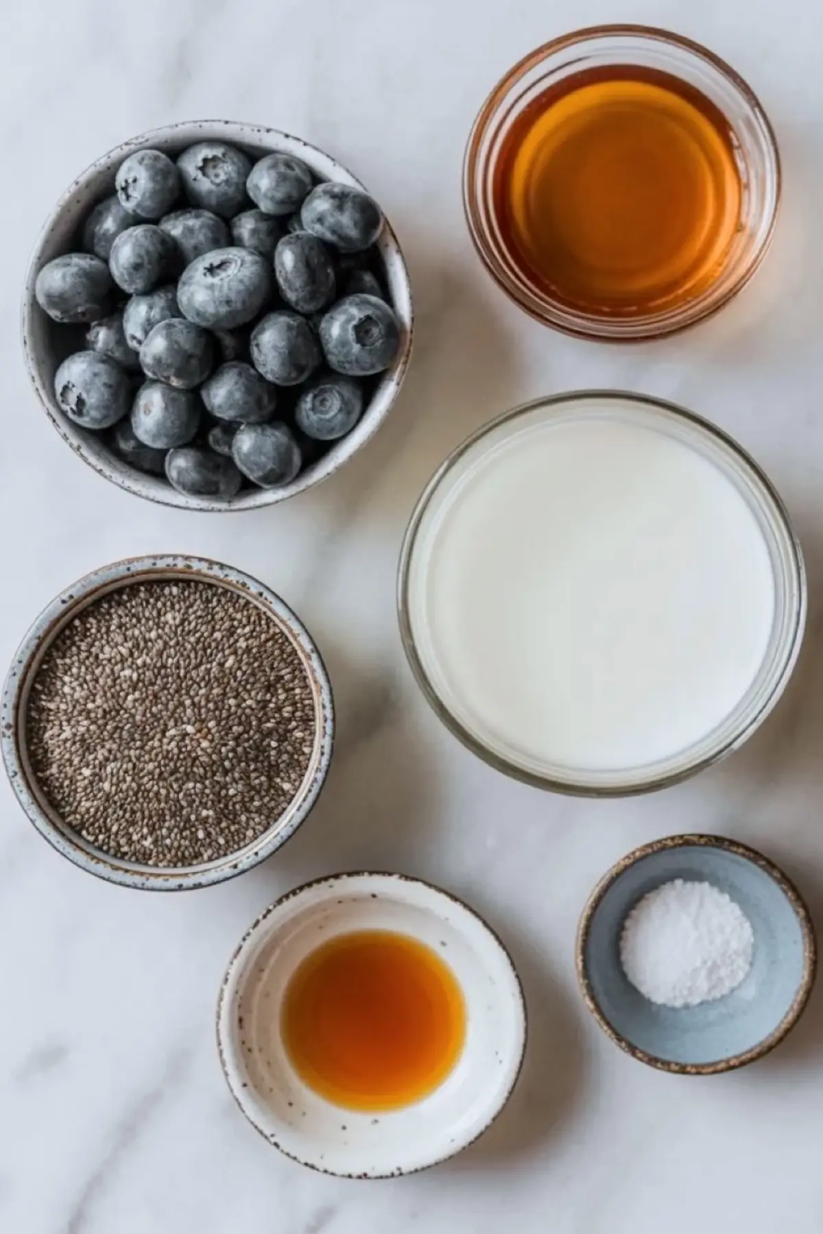 Overhead view shows fresh blueberries, chia seeds, milk, maple syrup, vanilla extract, and salt in small ceramic bowls on a white marble surface, creating a clean ingredient layout for blueberry chia seed pudding recipe and healthy breakfast prep.