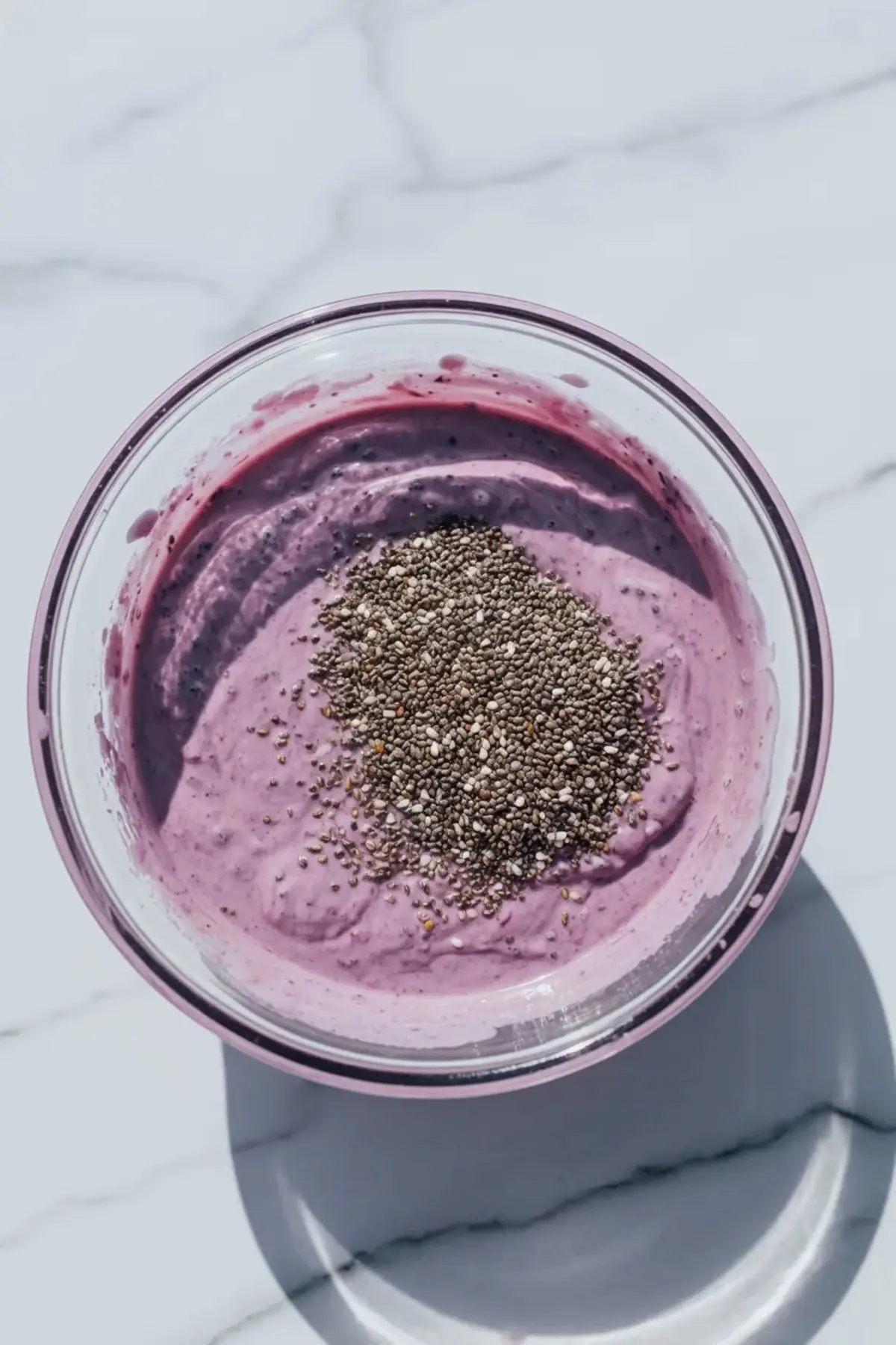 Overhead view shows blended blueberry smoothie mixture in a glass bowl topped with a mound of chia seeds on a marble surface, preparing blueberry chia seed pudding mixture for healthy breakfast meal prep.