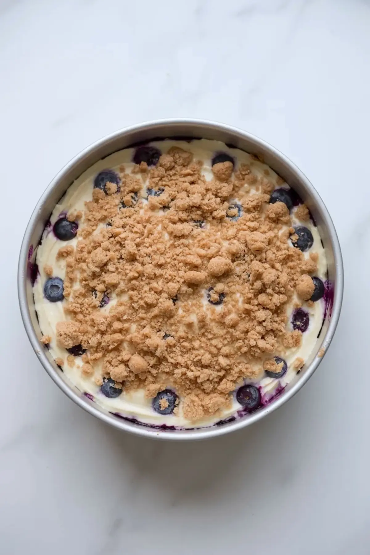 Round cake pan filled with thick vanilla cake batter topped with fresh blueberries and generous brown sugar crumb topping, overhead view on white marble surface, blueberry crumb cake before baking.
