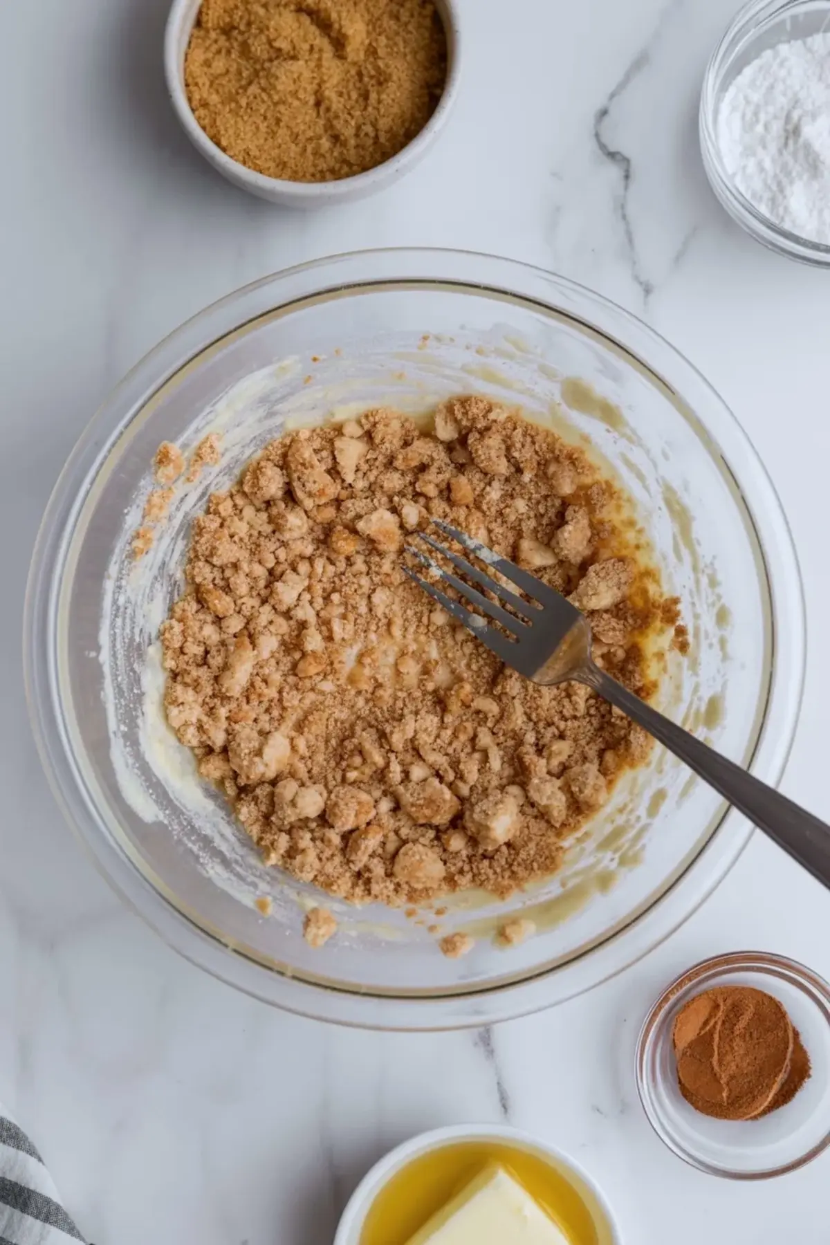 Glass mixing bowl contains brown sugar crumb mixture with melted butter and cinnamon while fork blends streusel topping, ingredients arranged on marble surface, homemade crumb cake topping preparation.
