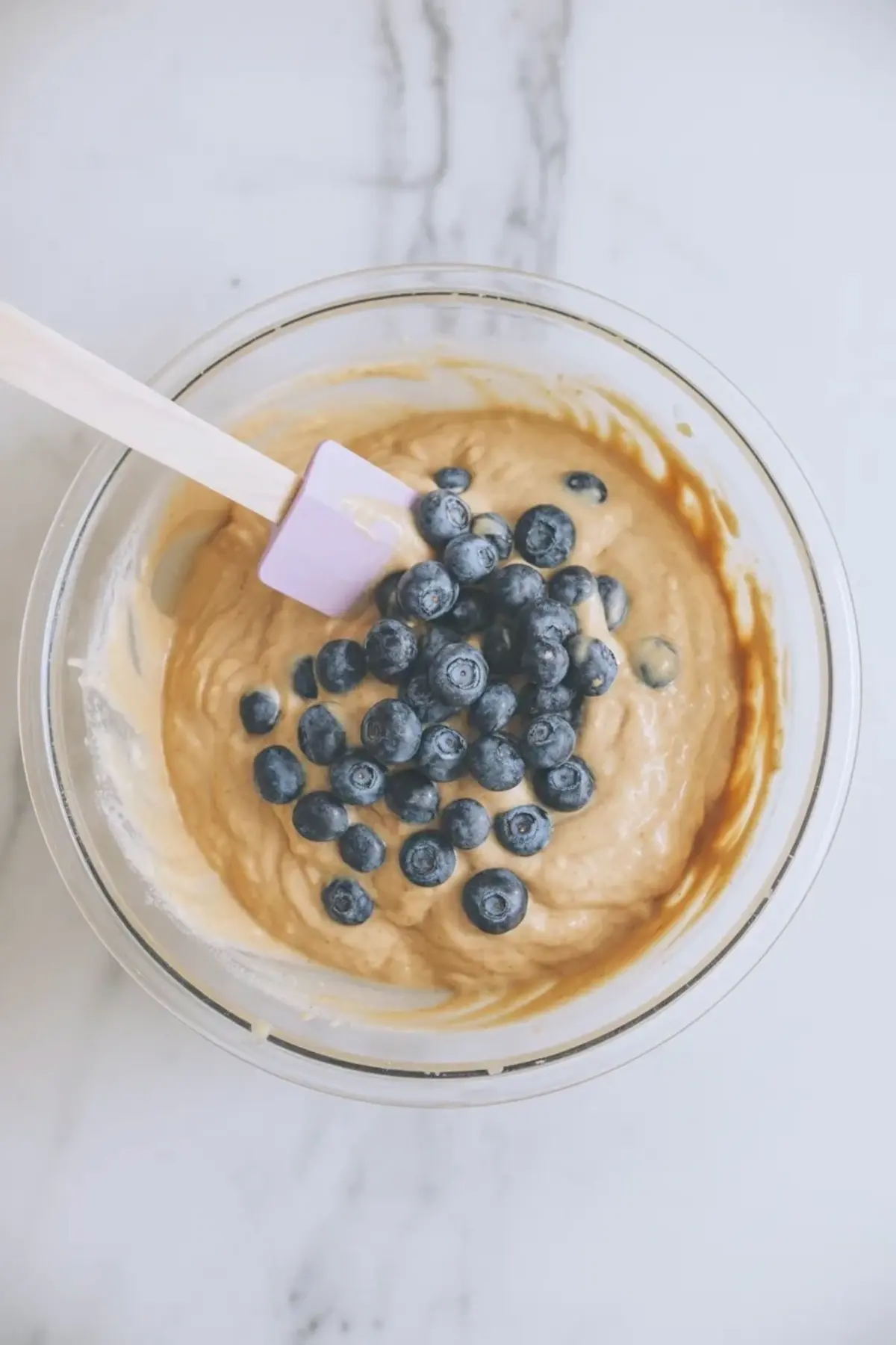 Fresh blueberries sit on thick batter inside a clear mixing bowl with a silicone spatula. Blueberries rest on creamy cottage cheese batter for a high protein baked breakfast recipe.
