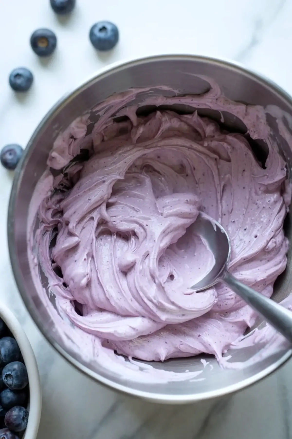 Bowl holds whipped blueberry cream cheese frosting with a smooth purple swirl and visible berry specks, while fresh blueberries sit on a white marble surface beside the bowl.