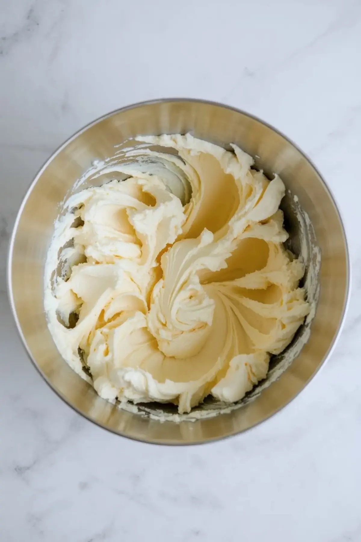 Metal mixing bowl contains creamed butter and sugar whipped into a pale yellow, fluffy mixture with soft peaks and smooth ridges on a light marble countertop.