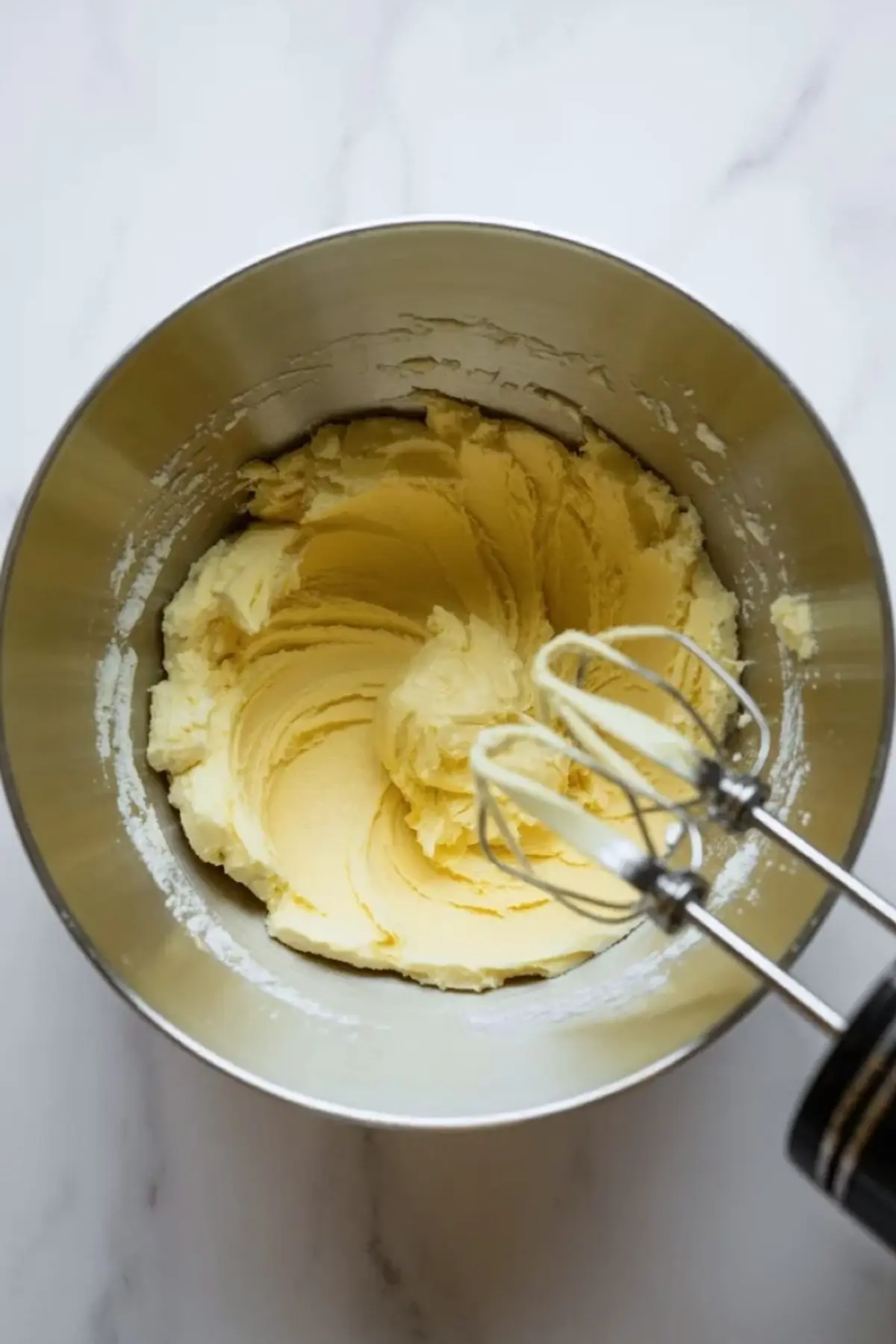 Electric hand mixer whips softened butter and sugar into a creamy pale yellow batter inside a metal mixing bowl, creating smooth spiral patterns along the sides.