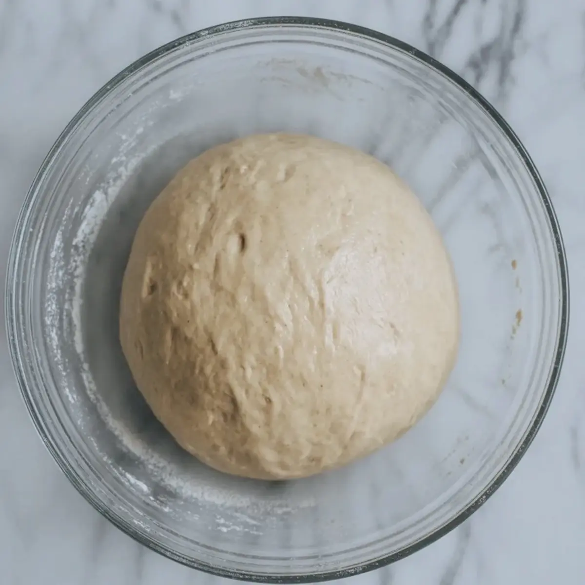 Smooth ball of enriched yeast dough rests in a clear glass bowl on a marble surface. The dough shows a soft texture and slight rise, ready for baking sweet cardamom bread, homemade brioche, or braided loaf recipe.
