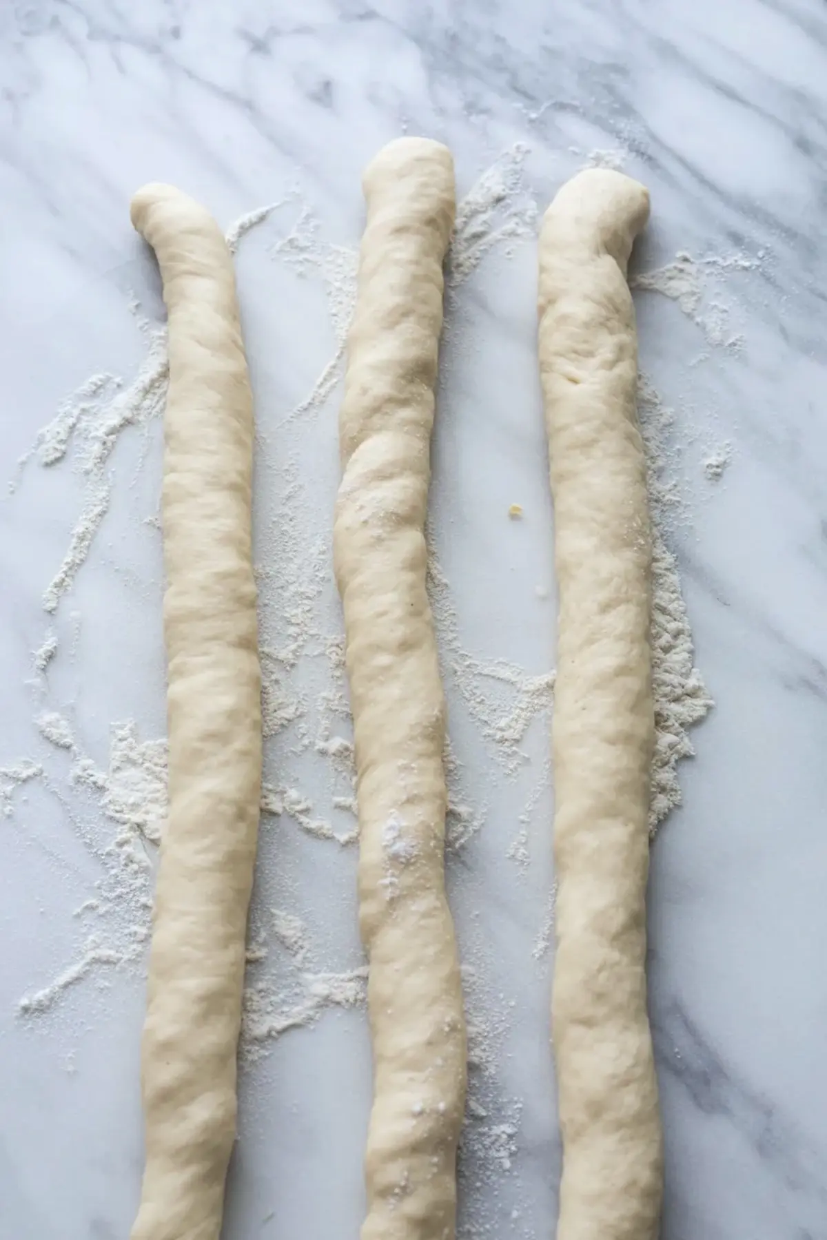 Three long ropes of yeast dough lie on a floured marble surface, shaped for braiding cardamom bread. The dough shows smooth texture and even thickness, prepared for homemade sweet braided loaf.
