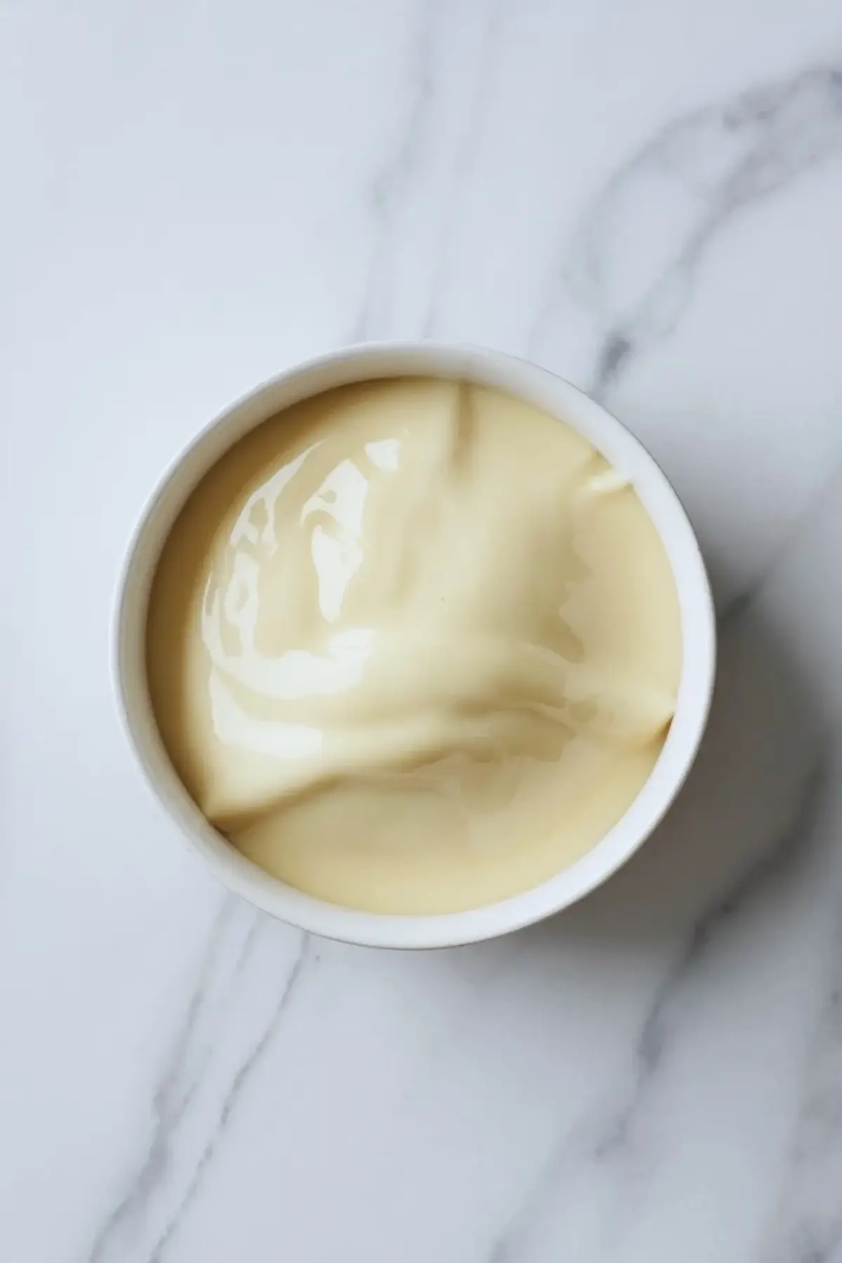 Smooth cream cheese frosting swirled in a white ceramic bowl placed on a light marble countertop.
