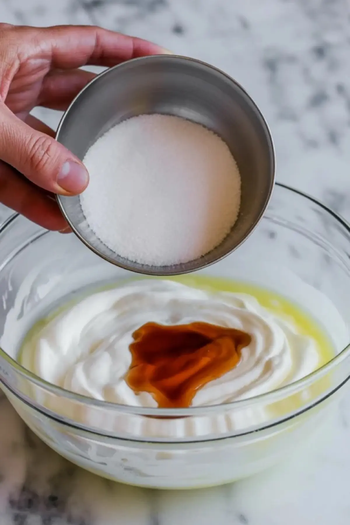 Hand holding a metal bowl of granulated sugar over a glass bowl containing whipped cream, vanilla extract, and yellow liquid ingredients on a marble surface.
