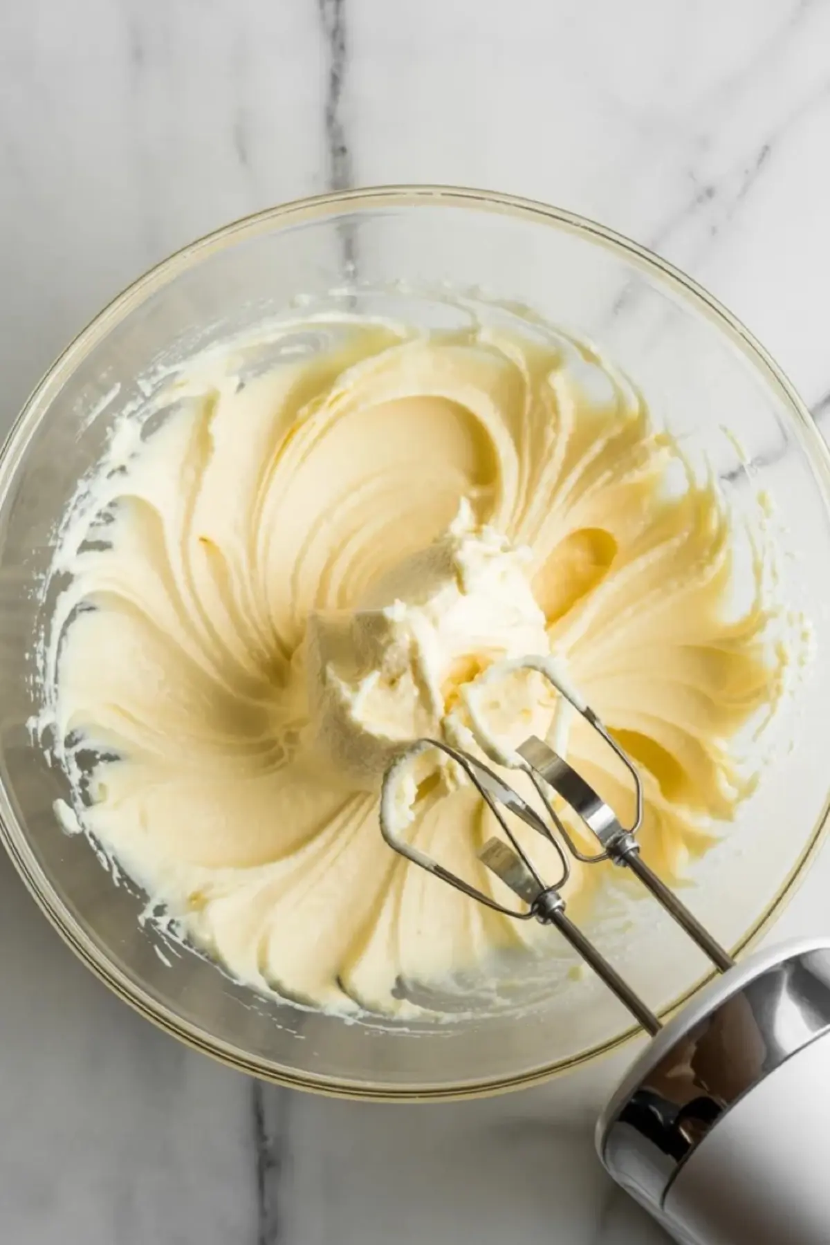 Electric hand mixer resting inside a glass bowl filled with thick whipped Chantilly frosting, with soft peaks swirled across the surface on a marble countertop.
