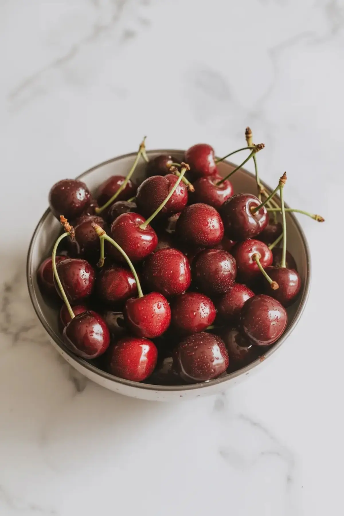 Bowl holds fresh whole cherries with stems and water droplets on a light marble background. The ripe red cherries display natural shine and deep color.