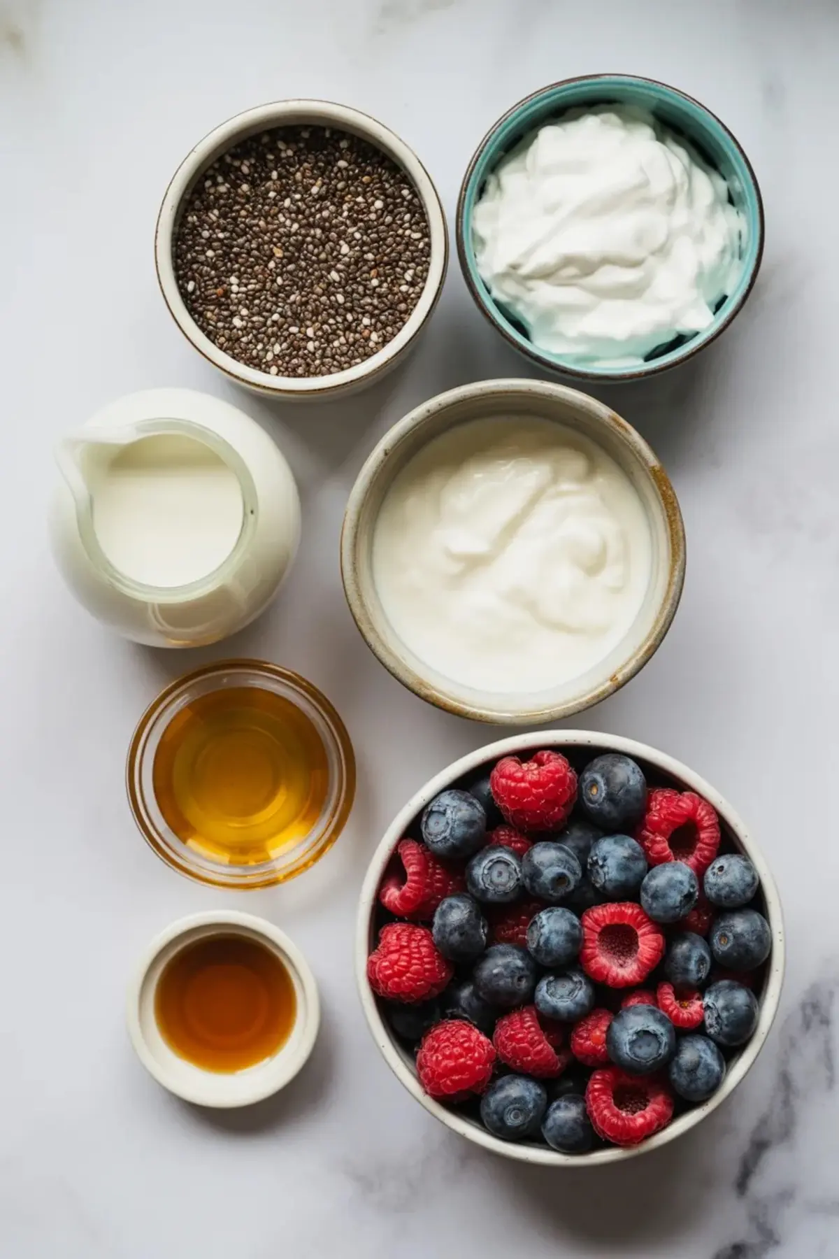 Small bowls contain chia seeds, Greek yogurt, milk, honey, and vanilla extract beside fresh blueberries and raspberries on a white marble surface. Ingredients prepare a healthy chia seed pudding with yogurt and berries.
