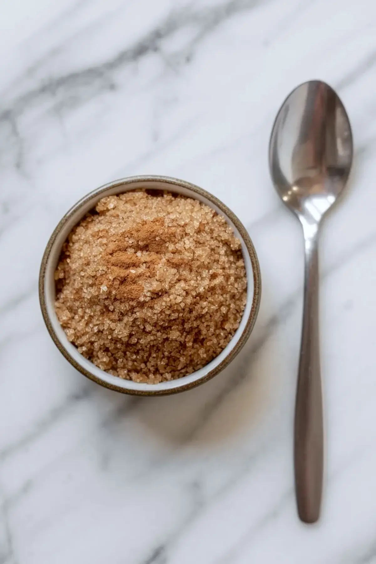 Small ceramic bowl filled with coarse brown sugar topped with ground cinnamon, placed beside a metal spoon on a white marble background.