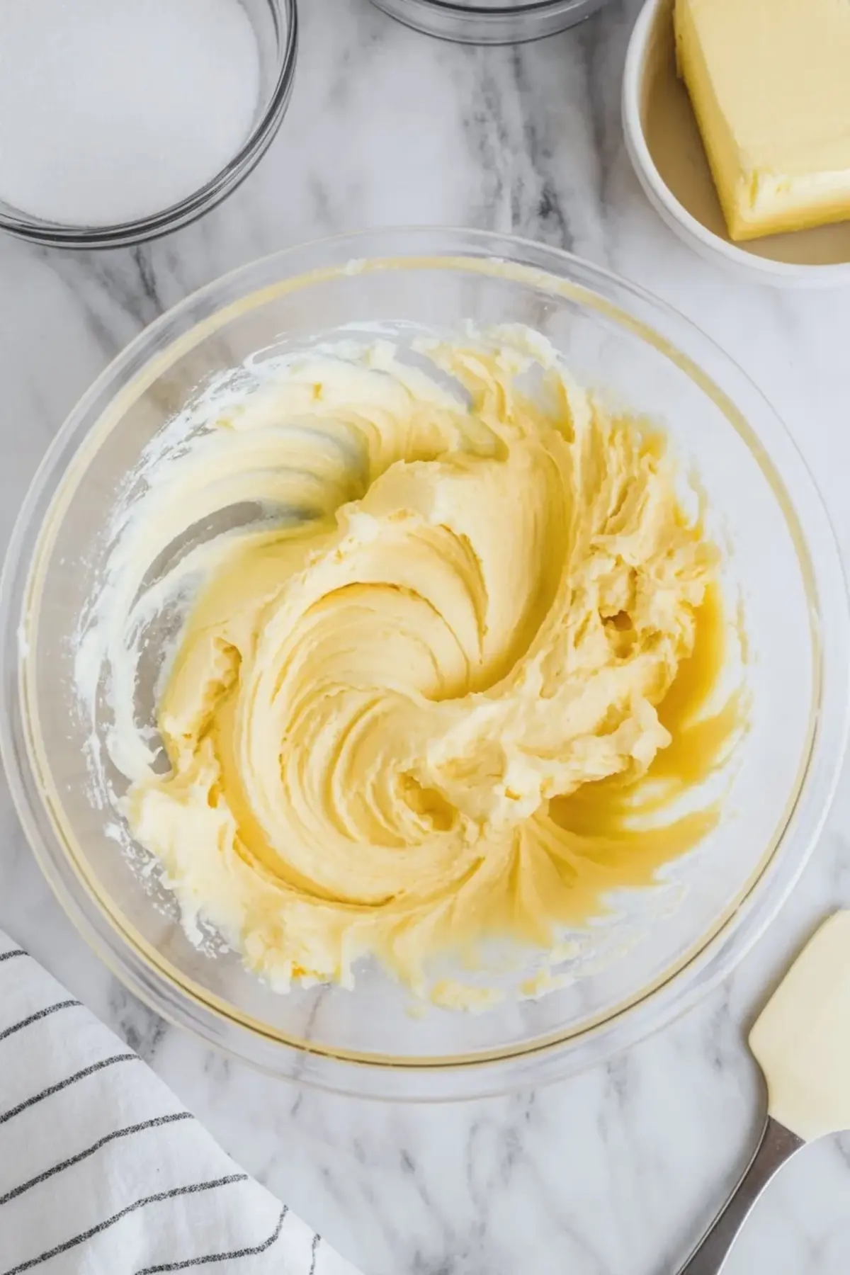 Overhead shot of whipped butter and sugar in a glass bowl with surrounding ingredients including granulated sugar and a block of butter on a marble background.