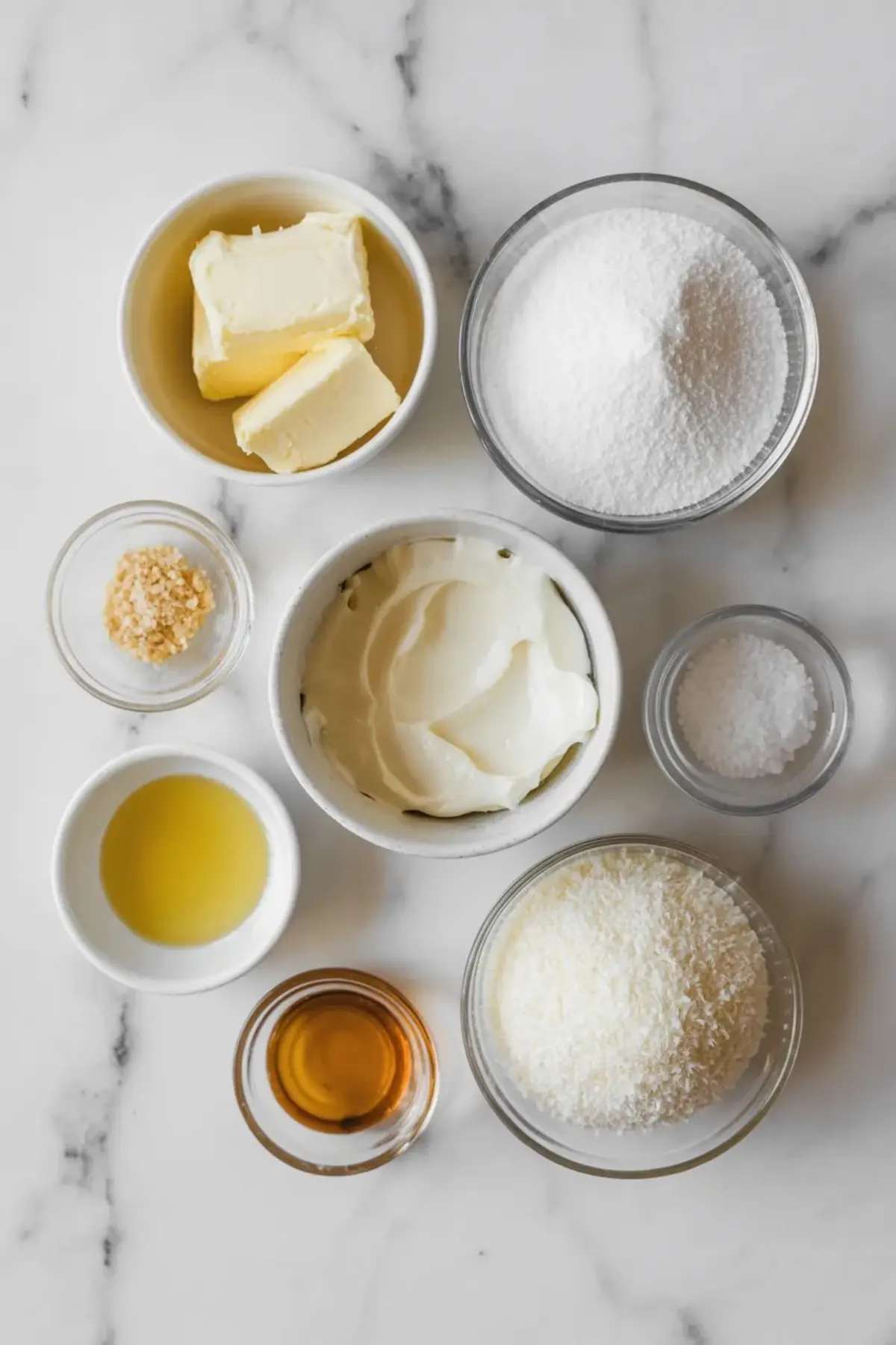 Small bowls display butter, powdered sugar, cream cheese, shredded coconut, vanilla extract, lemon juice, and salt on a marble countertop. Ingredients are arranged flat lay style for coconut buttercream frosting recipe prep.