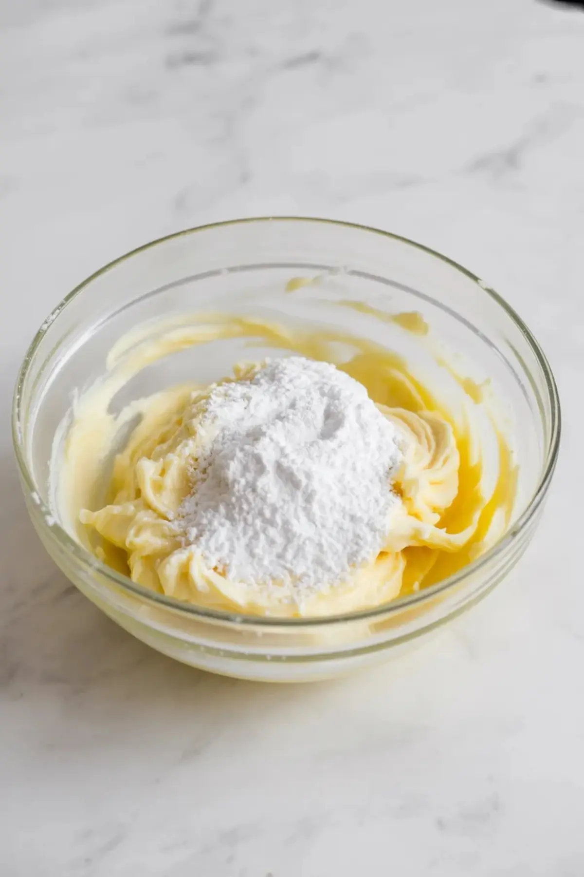 Glass bowl contains whipped butter mixture topped with a mound of powdered sugar. The mixture shows the step of adding sugar for smooth coconut buttercream frosting.