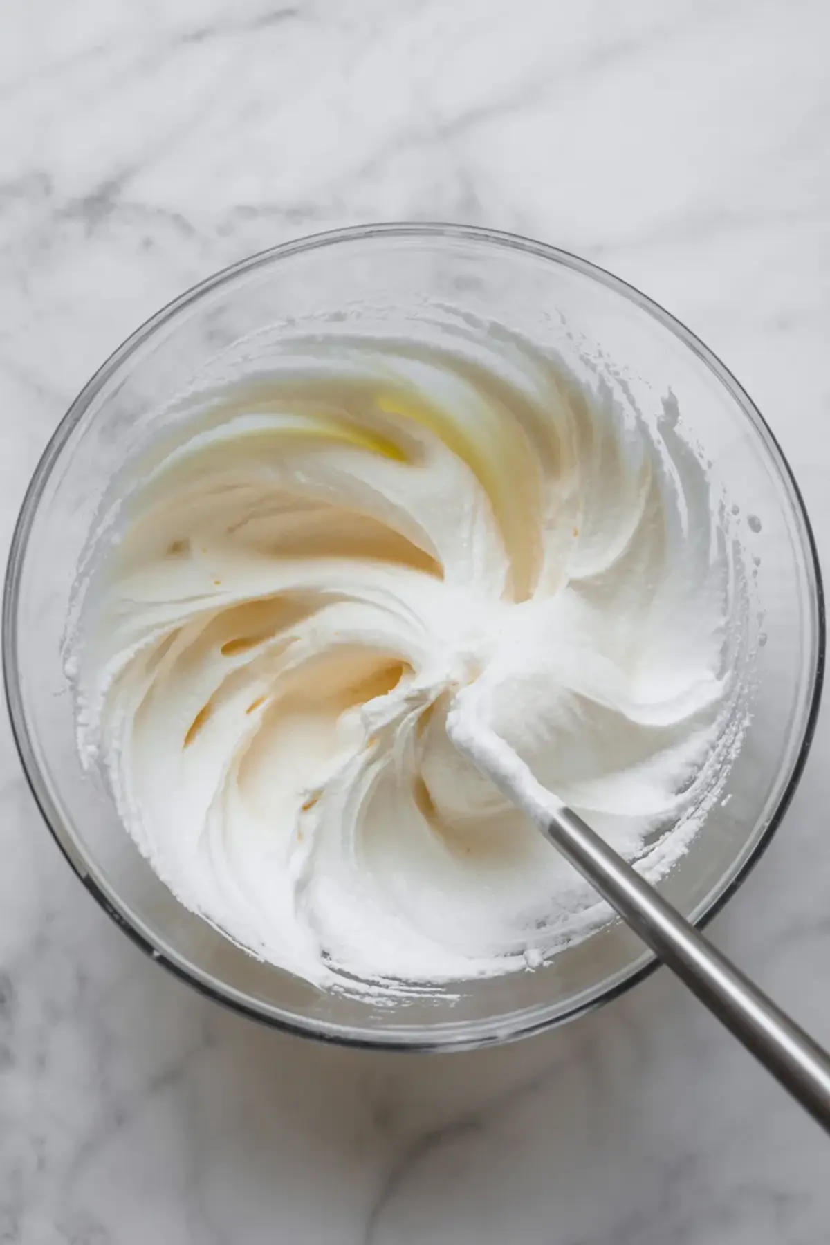 Glass bowl contains whipped coconut cream with visible powdered sugar and vanilla streaks. Metal whisk mixes the ingredients into a fluffy texture. The cream forms thicker swirls and soft peaks. This image shows how to whip coconut cream for cake filling and cupcake frosting.
