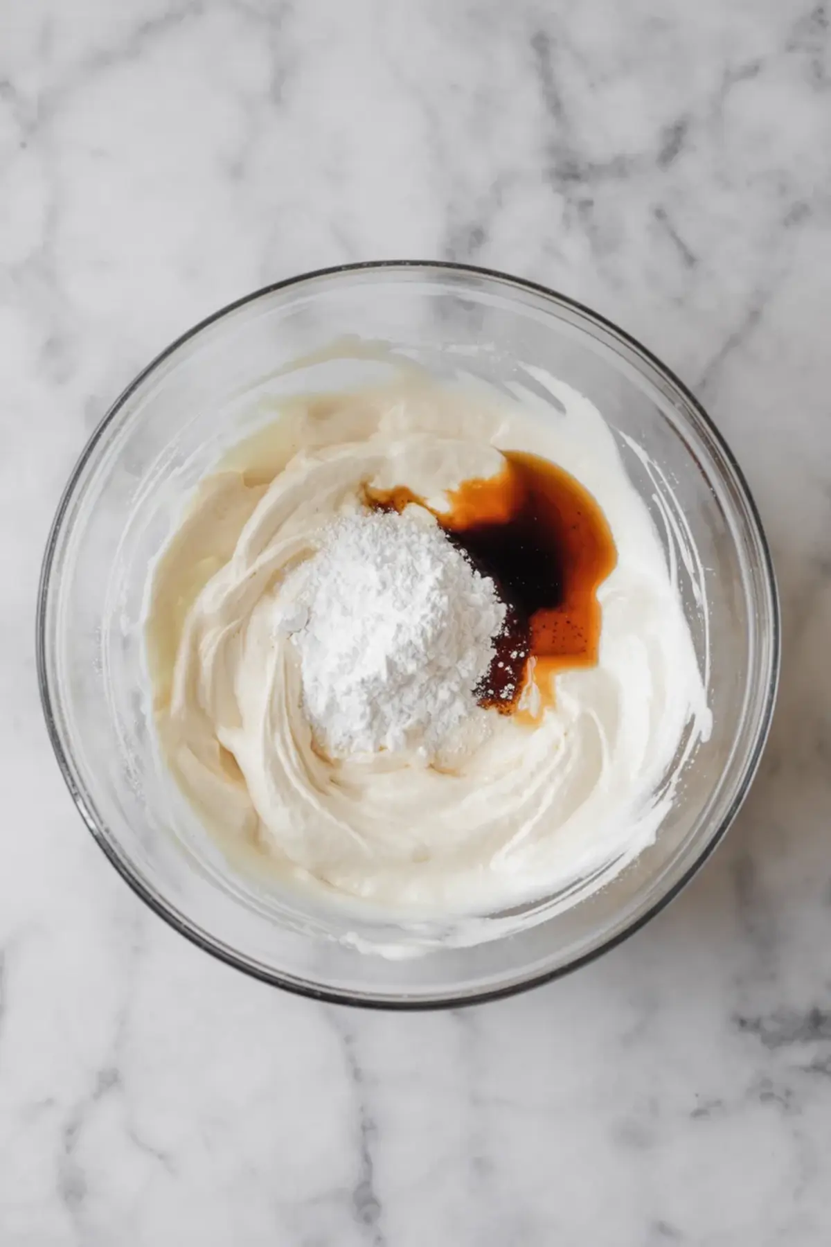 Glass bowl holds coconut cream with powdered sugar and vanilla extract added on top. The white sugar mound and brown vanilla contrast with the creamy base. This step shows ingredients for homemade coconut whipped cream before mixing for vegan baking and dairy free desserts.
