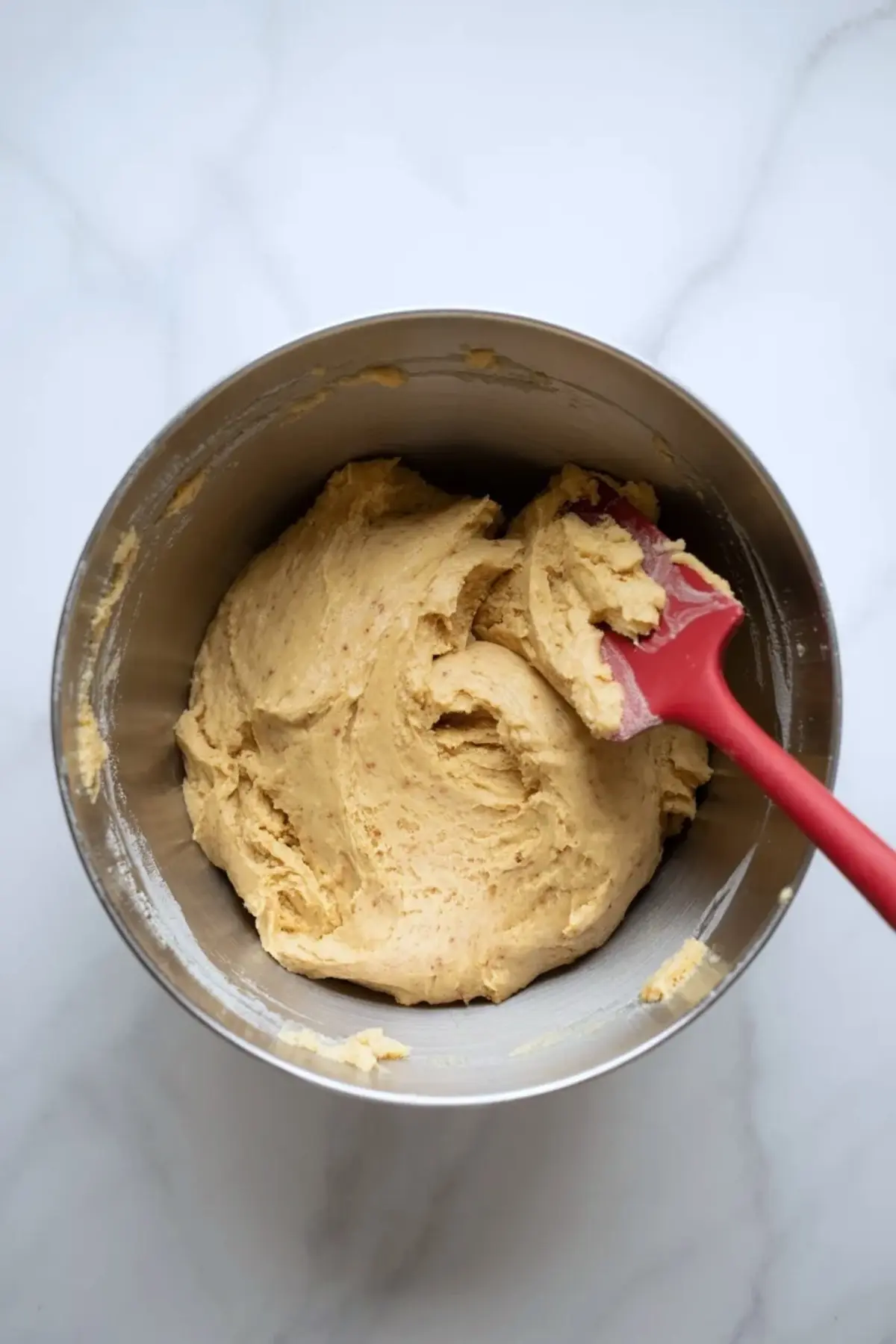 Metal mixing bowl holds thick vanilla cookie dough with red spatula on marble countertop, showing soft dough texture for homemade coffee cake cookies.