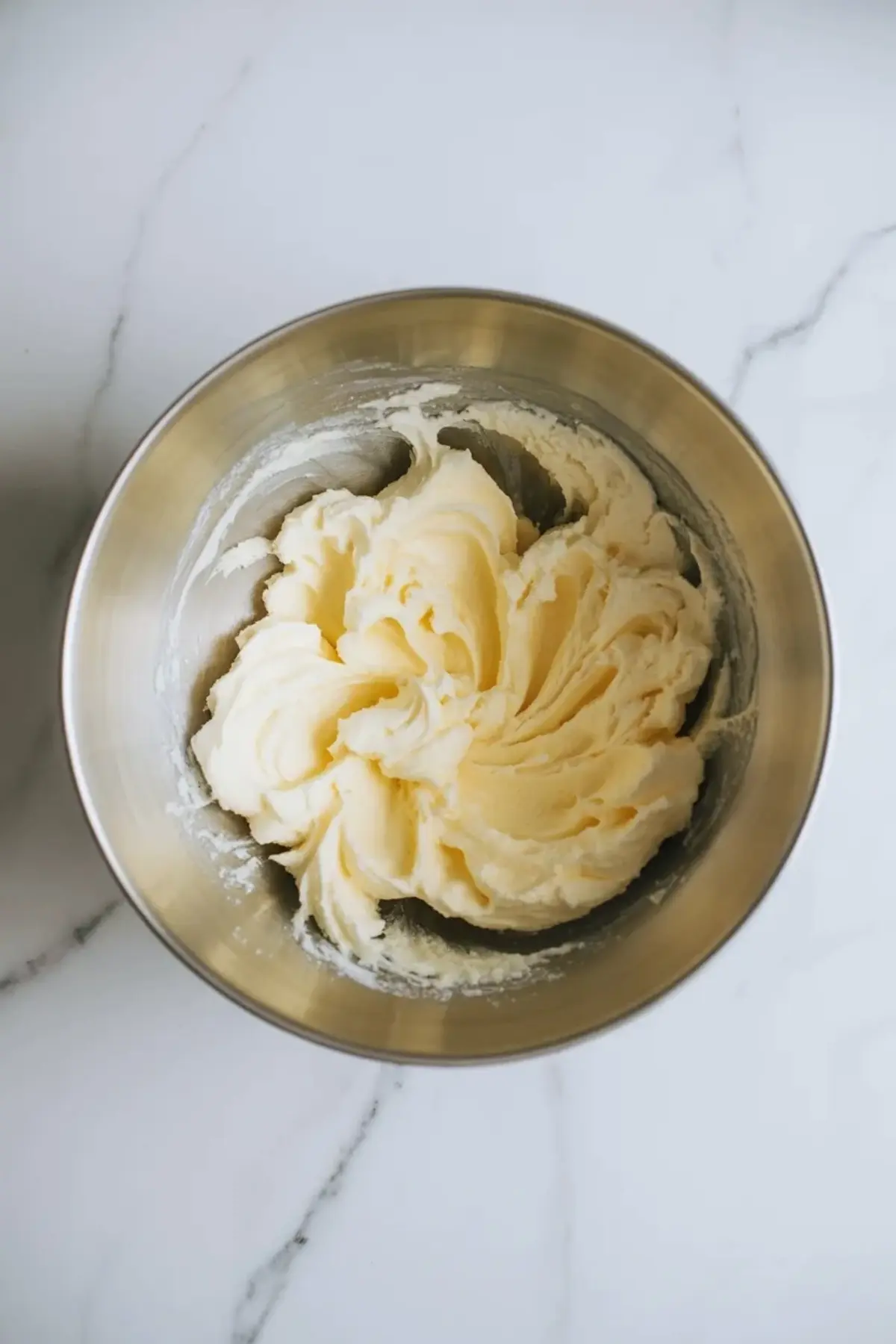 Metal bowl contains creamed butter and sugar mixture with fluffy pale texture, prepared for coffee cake cookie batter on marble background.