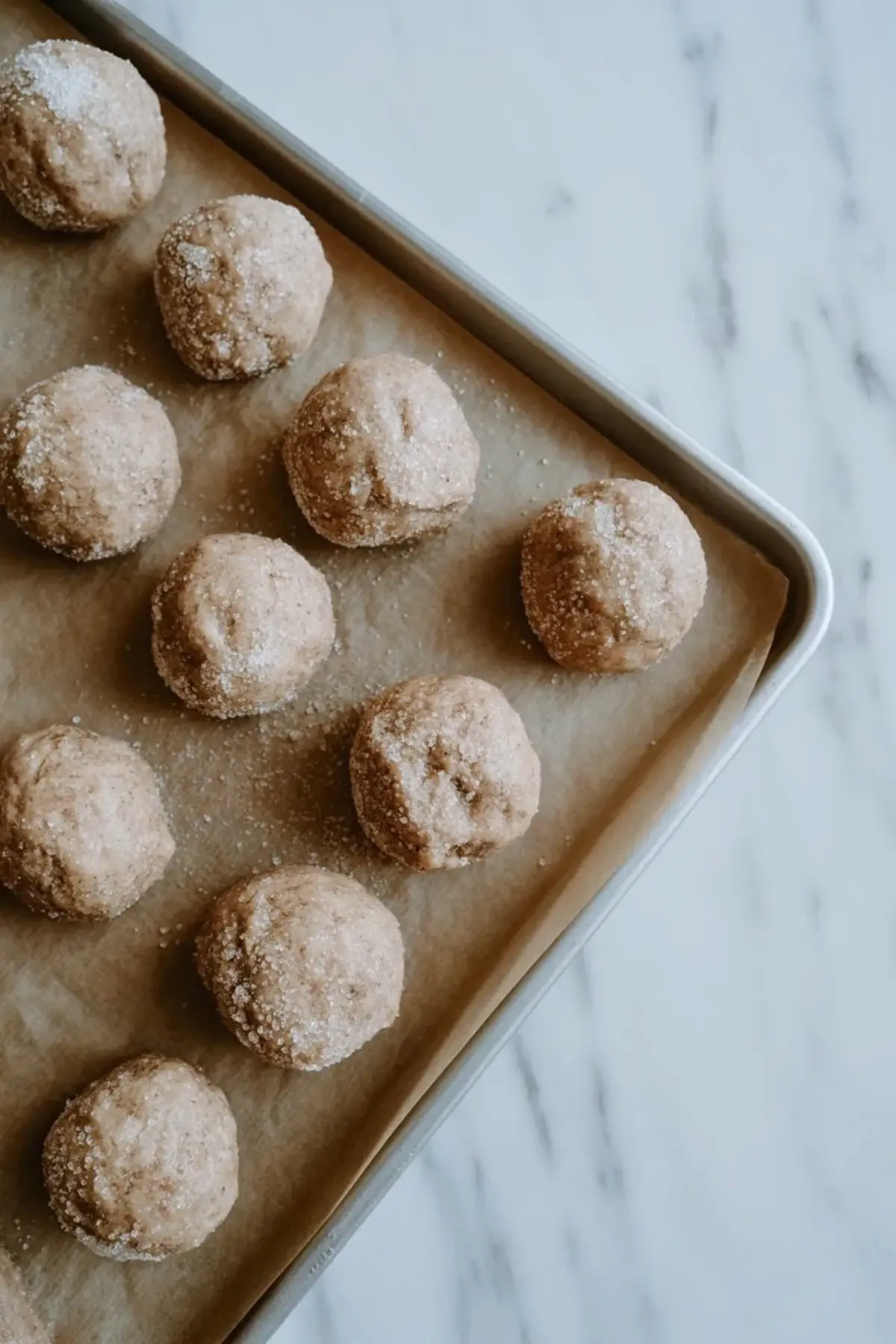 Round cookie dough balls coated in granulated sugar sit on a parchment lined baking sheet. Even scoops of cardamom sugar cookie dough show light sugar coating and smooth texture. Overhead view shows homemade cookie dough ready for baking.