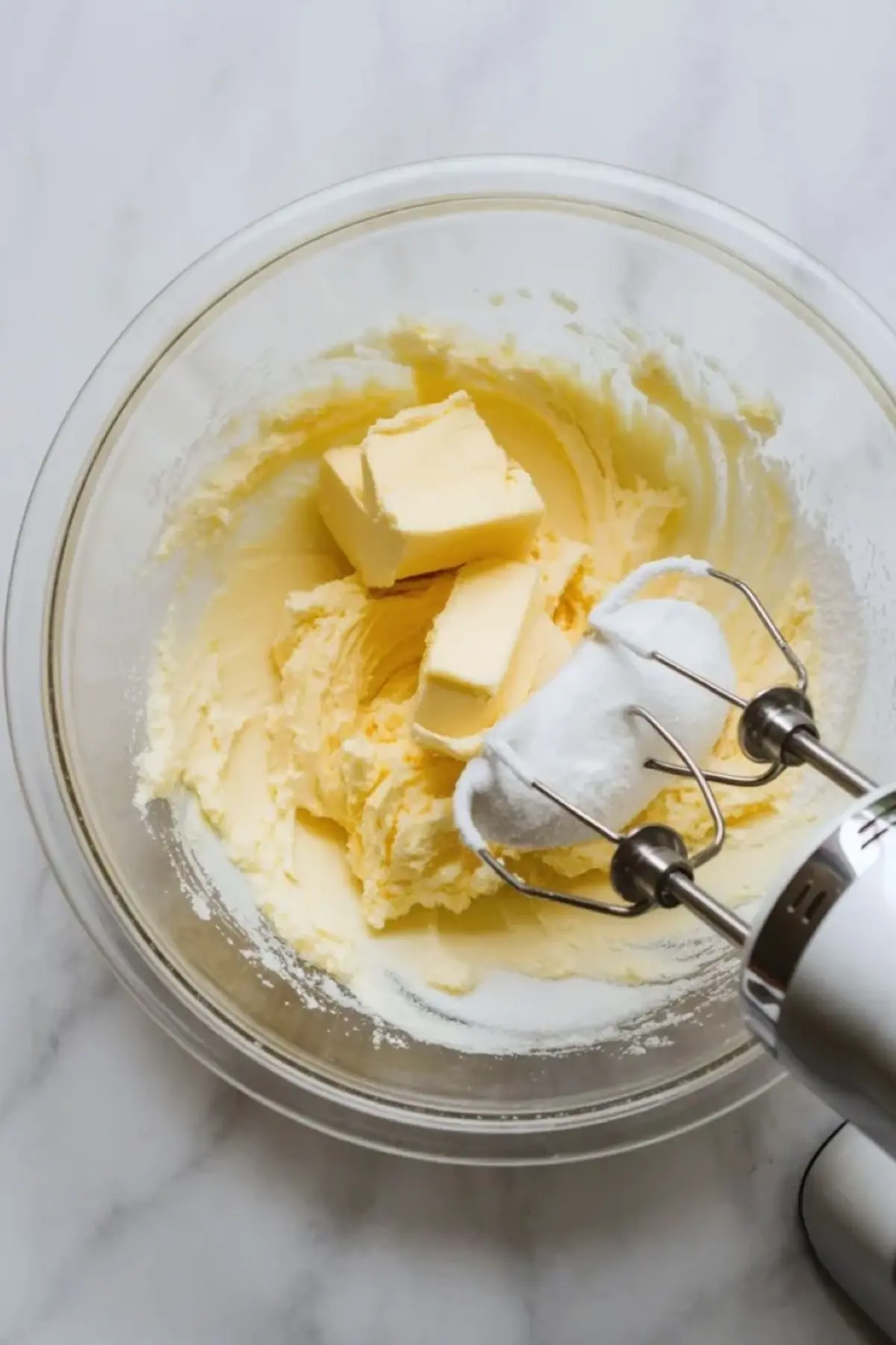 Hand mixer blends softened butter and granulated sugar in a clear glass bowl. Thick and creamy butter mixture clings to metal beaters. Close up shows creaming butter and sugar for coffee cardamom sugar cookie dough.