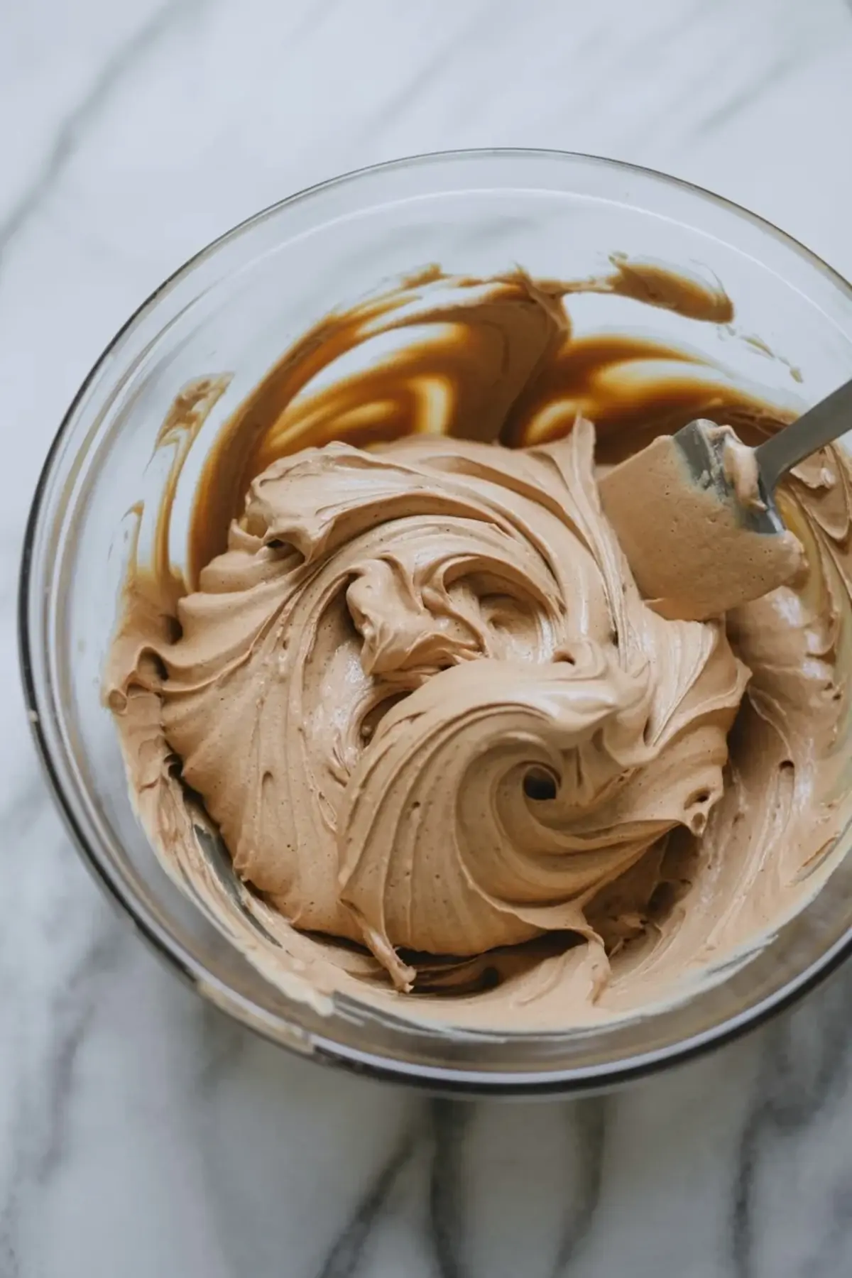 Light brown coffee buttercream frosting swirls in a clear glass bowl with a spatula. Creamy espresso frosting shows a smooth texture and glossy finish for topping coffee cupcakes.