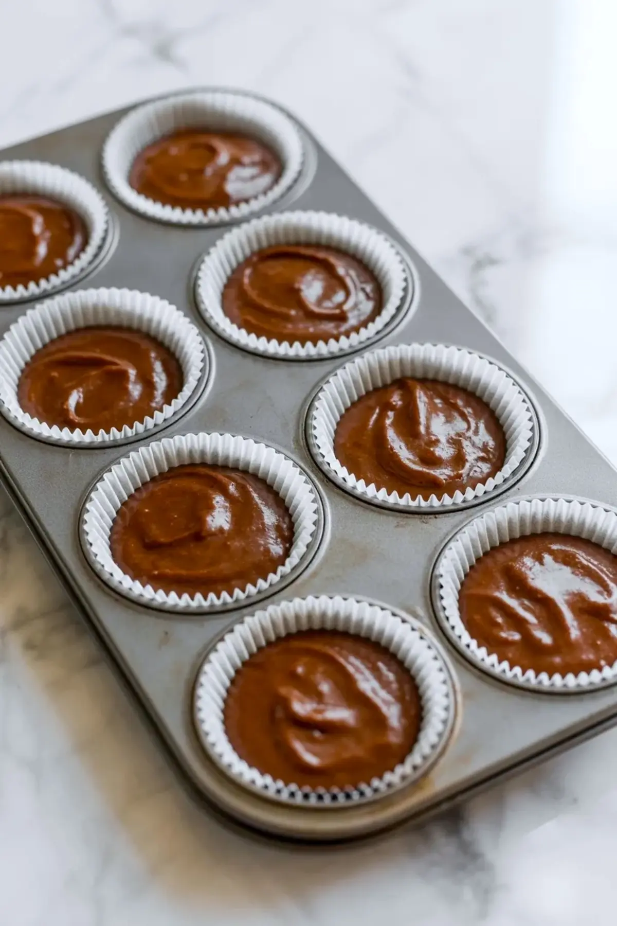 Chocolate cupcake batter fills white paper liners in a metal muffin tin on a marble countertop. Smooth coffee chocolate batter sits evenly in each cup, ready for baking homemade mocha cupcakes.