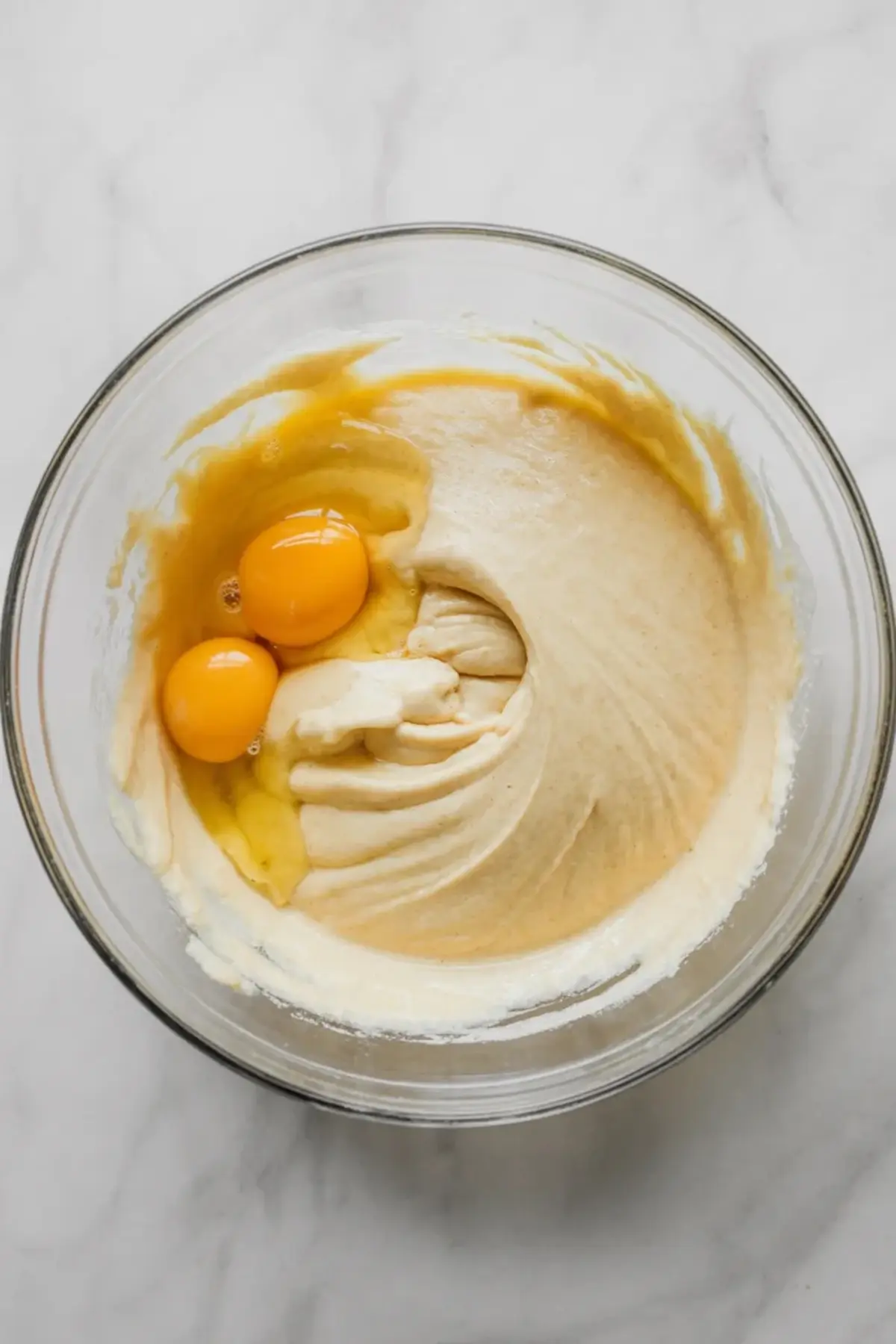 Two raw eggs sit in a bowl of creamed butter and sugar. Yellow egg yolks rest on smooth cake batter as ingredients combine for homemade coffee cupcake recipe.