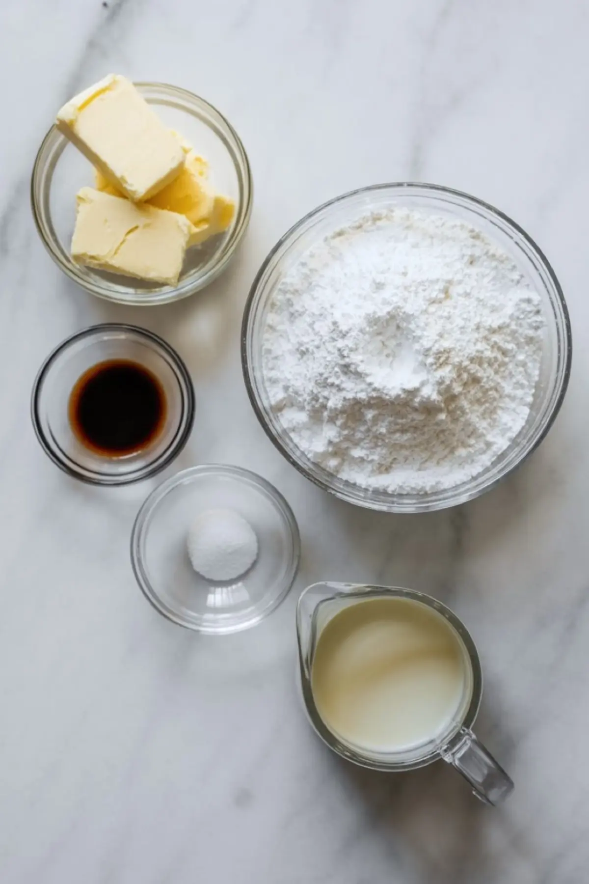 Measured ingredients sit on marble surface including softened butter cubes, powdered sugar in glass bowl, vanilla extract, baking powder, and milk in measuring cup for cookie frosting recipe.

