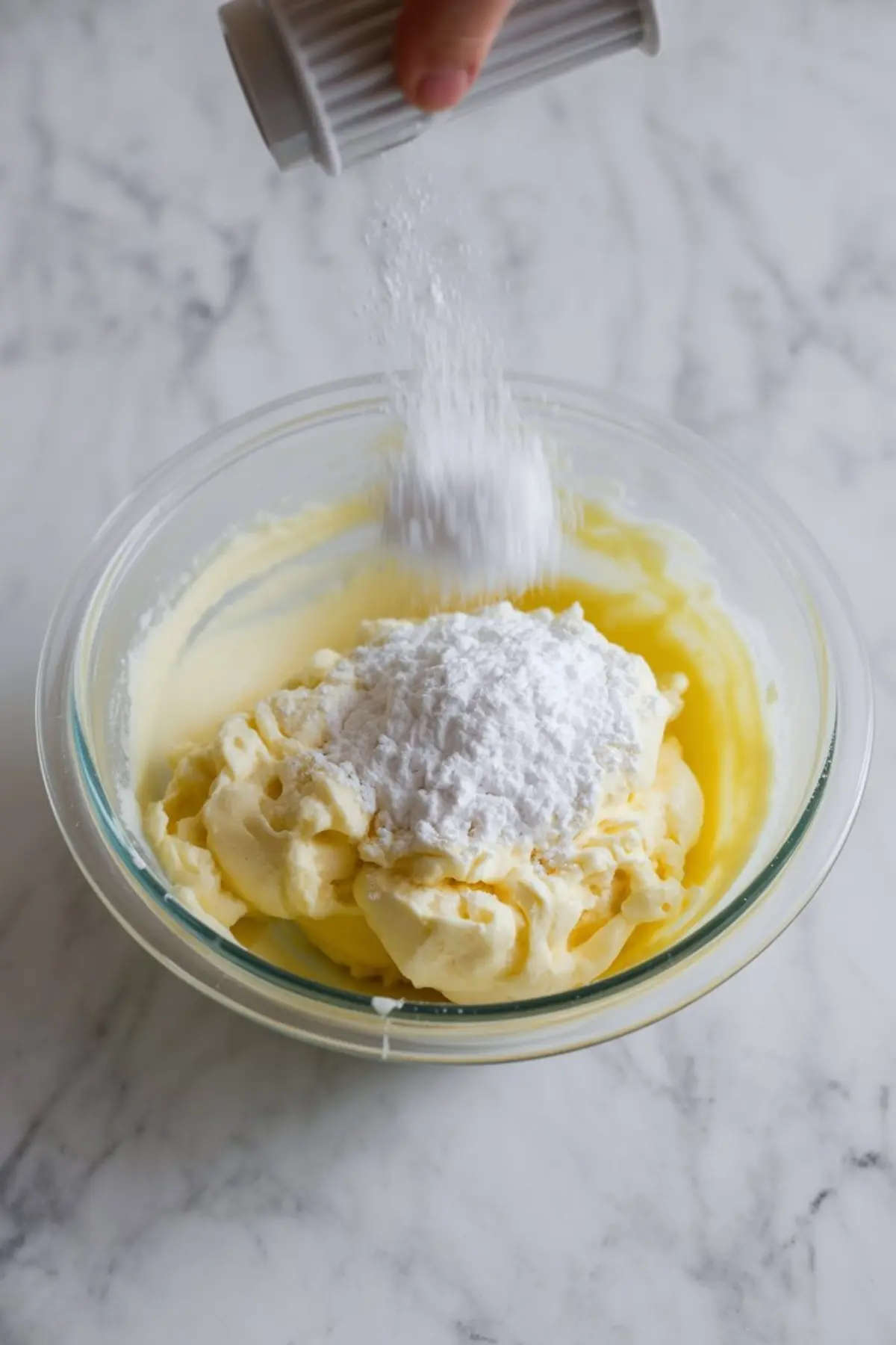 Hand sprinkles powdered sugar into bowl of creamed butter mixture. White sugar falls over pale yellow frosting base in glass bowl, step of mixing homemade buttercream frosting.
